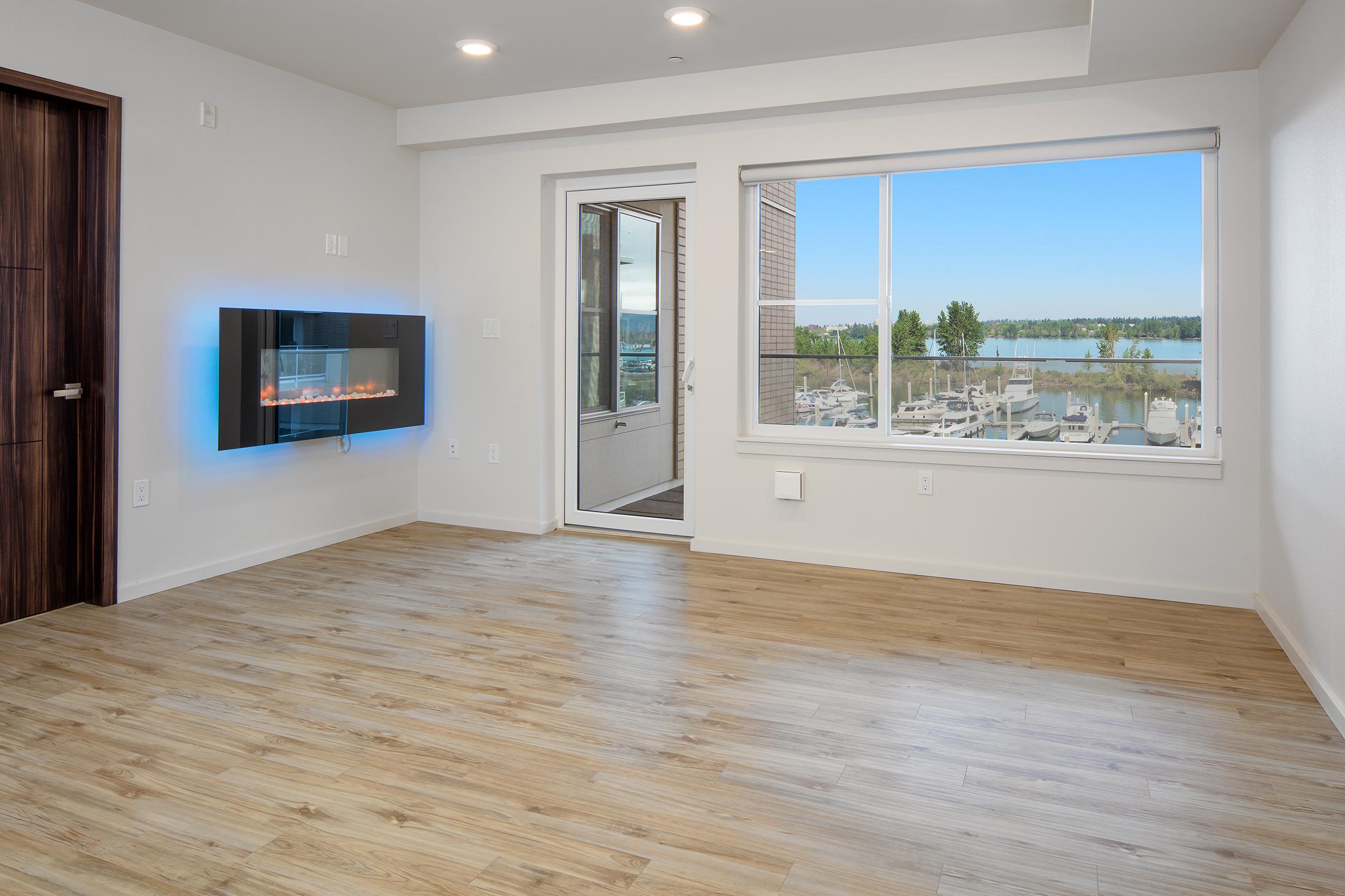 A modern, spacious living room featuring light wood flooring, a wall-mounted electric fireplace, large windows with a lake view, and a sliding glass door leading to a balcony. The walls are painted light neutral colors, enhancing the bright and airy atmosphere of the space.