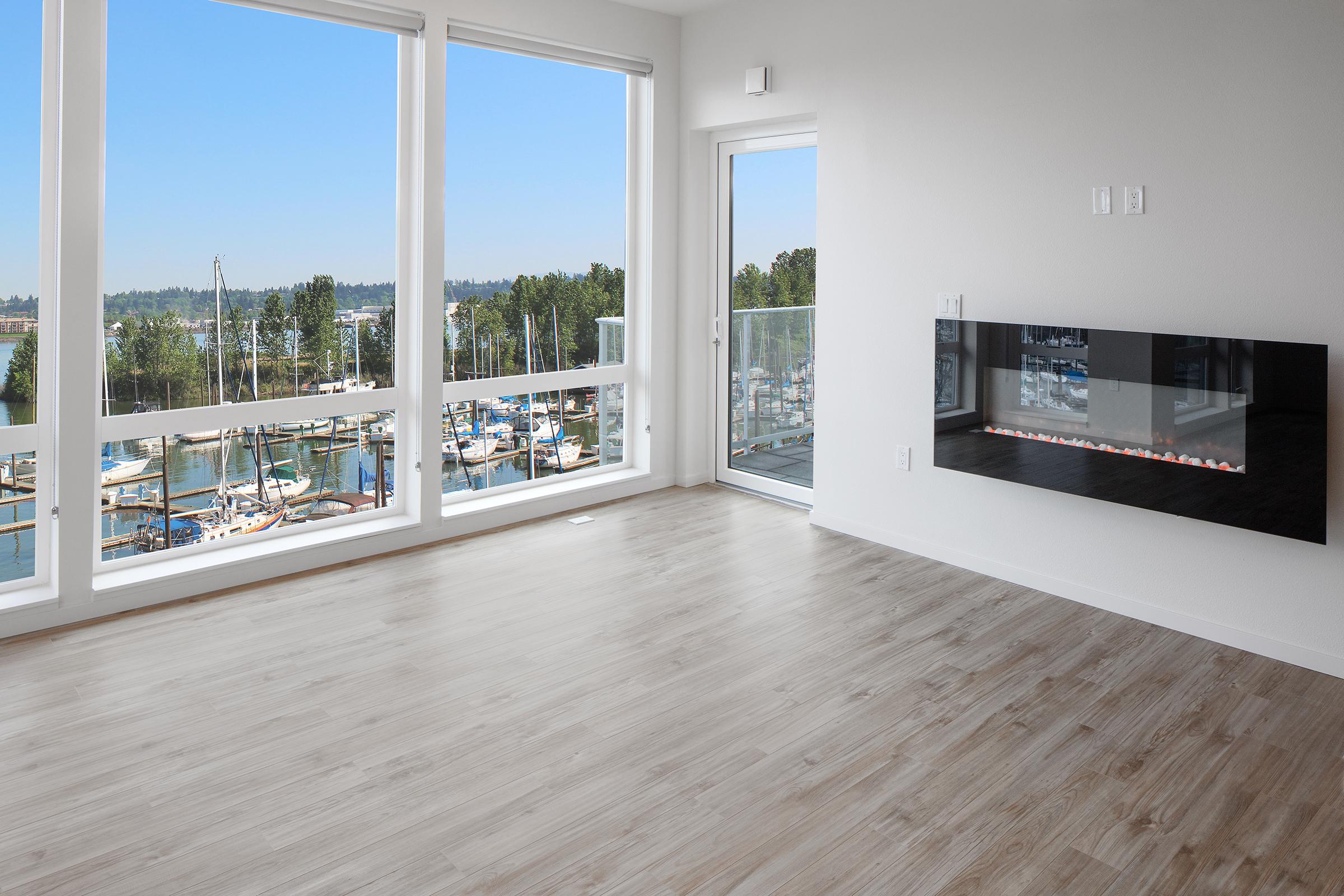 A modern, minimalistic living room with large windows showcasing a view of a marina. The space features light wood flooring, a built-in wall-mounted fireplace, and access to a balcony. Bright natural light floods the room, creating a welcoming atmosphere.