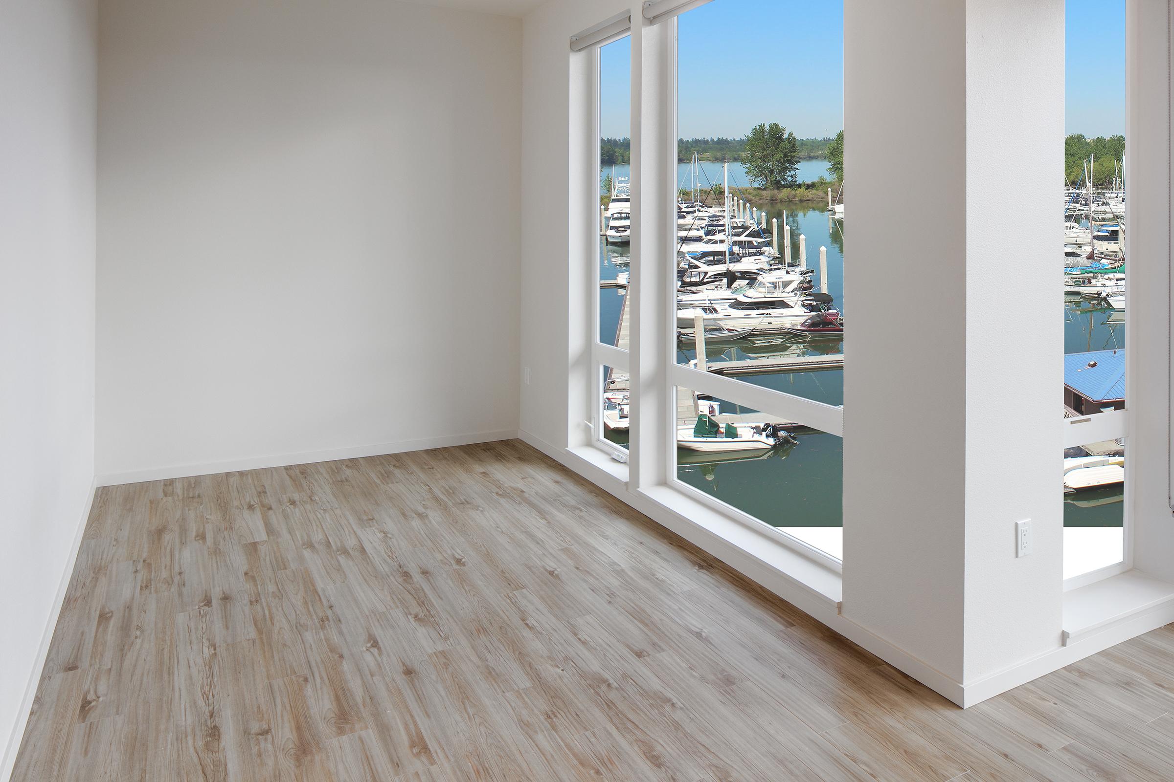A bright, empty room with light brown wooden flooring and large windows showcasing a marina filled with boats and a clear blue sky. The walls are painted white, creating a spacious and airy feel.