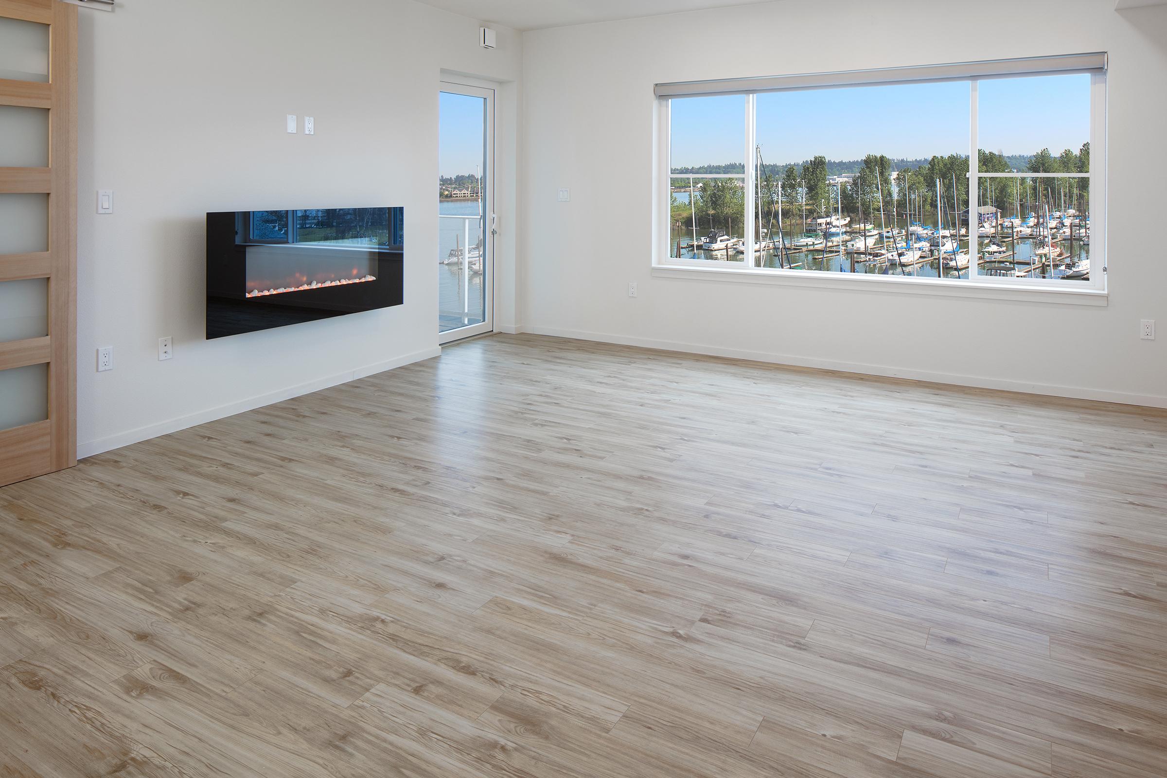 A bright, spacious room with light wood flooring. A large window offers a view of a marina with boats. On one wall, there is a modern black fireplace. The room has neutral-colored walls and an open door leading to a balcony, enhancing the airy feel.