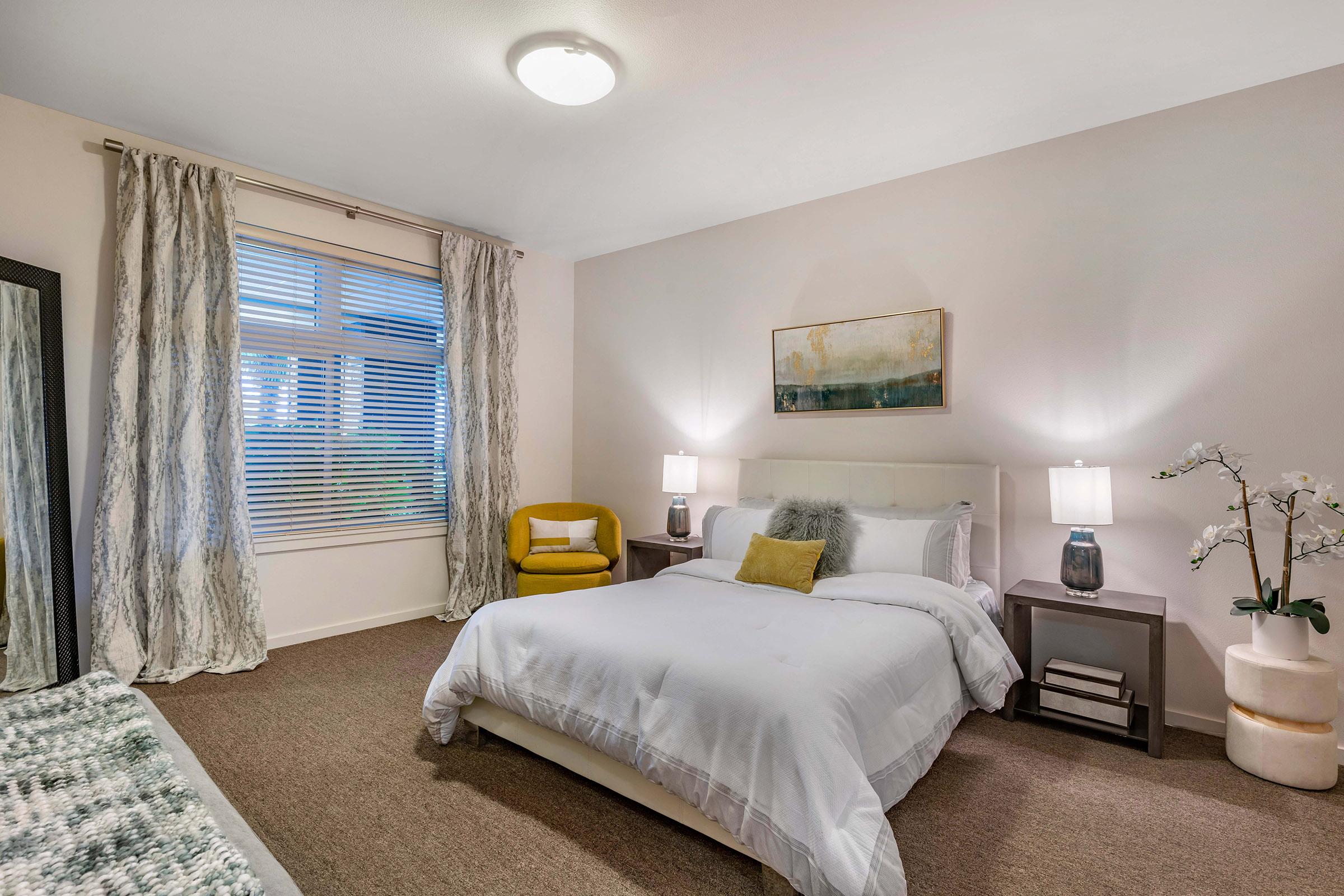A well-decorated bedroom featuring a white bed with gray and white bedding, two bedside tables with lamps, a yellow accent chair, and a large mirror. Light-colored curtains frame the window, allowing natural light to enter, while an abstract painting adorns the wall above the bed.