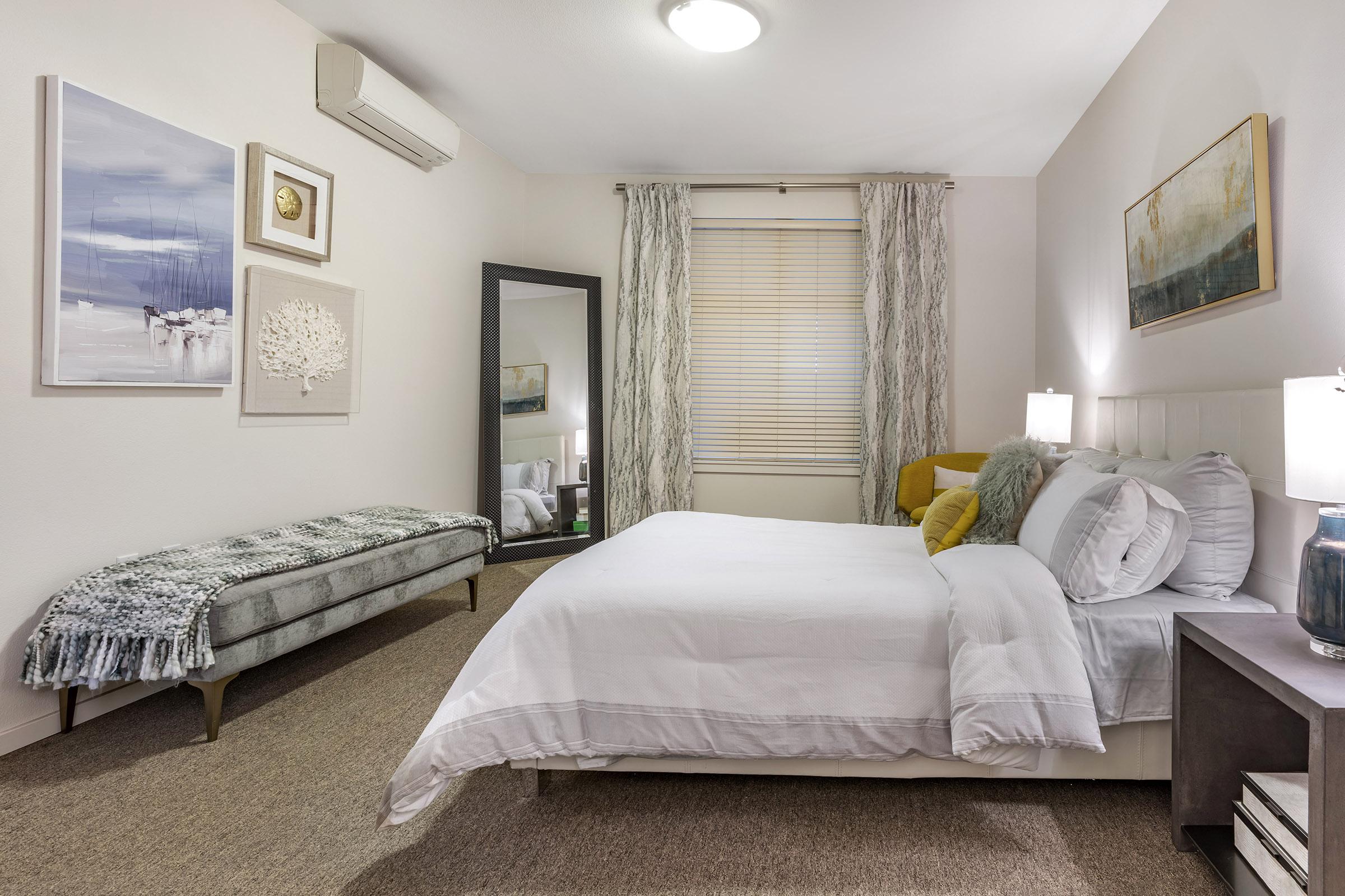 A cozy bedroom featuring a white bed with gray and yellow accents, a large mirror, and a stylish bench. The walls are adorned with contemporary artwork, and there are light-colored curtains. Soft lighting creates a warm atmosphere, enhancing the inviting decor of the space.