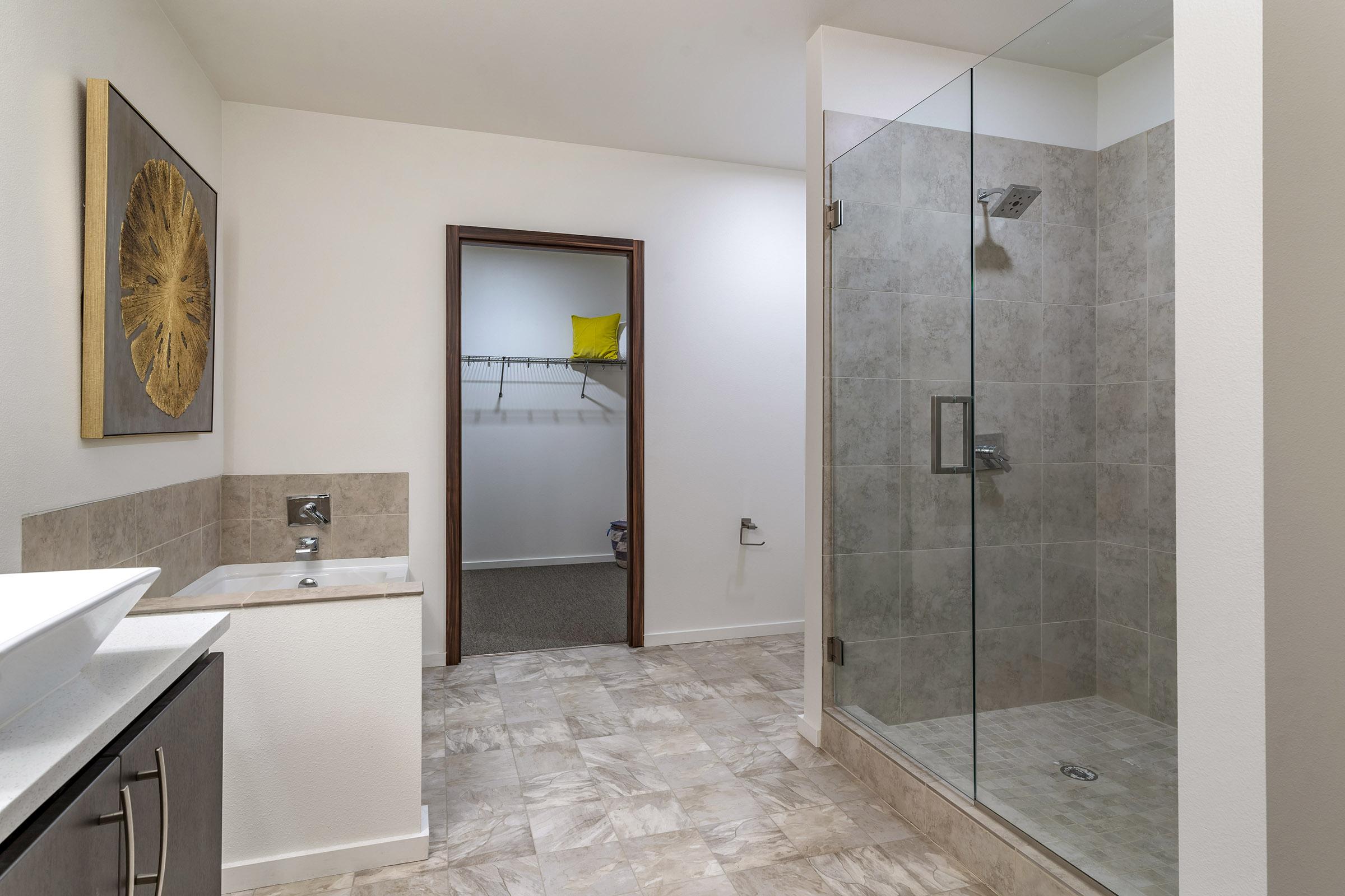 Modern bathroom featuring a spacious walk-in shower with glass door, a stylish sink with a countertop, and tiled walls. A doorway leads to a closet space, and a decorative piece is displayed on the wall. Neutral color scheme with light-colored flooring and walls.