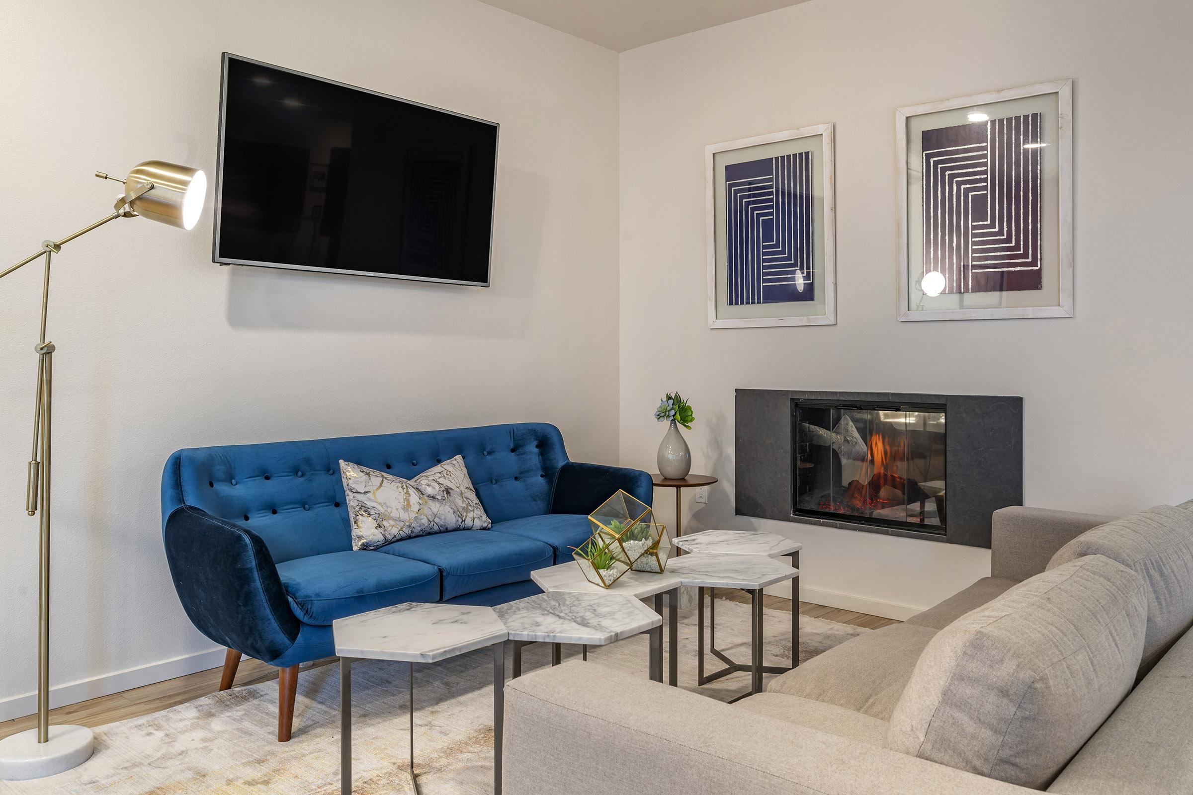 A modern living room featuring a blue velvet sofa with decorative pillows, a light gray sectional sofa, stylish marble-top side tables, a contemporary floor lamp, and a wall-mounted television. Above the fireplace, there are two framed abstract artworks, with a cozy fire burning in the fireplace.