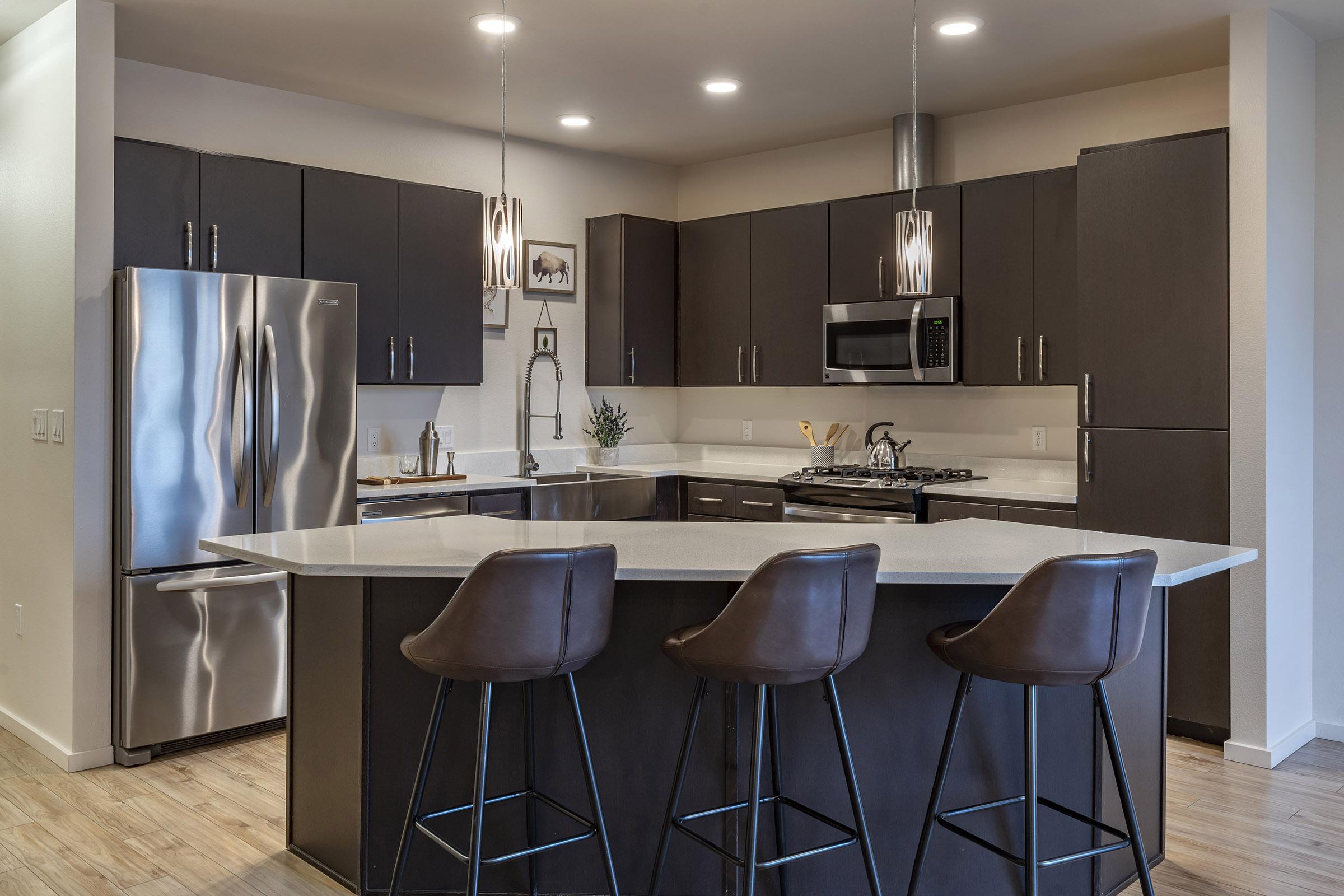 Modern kitchen featuring dark cabinets, stainless steel appliances, and a large island with three high bar stools. The space is bright, with recessed lighting and a contemporary design that includes a gas stove, microwave, and sleek countertops.