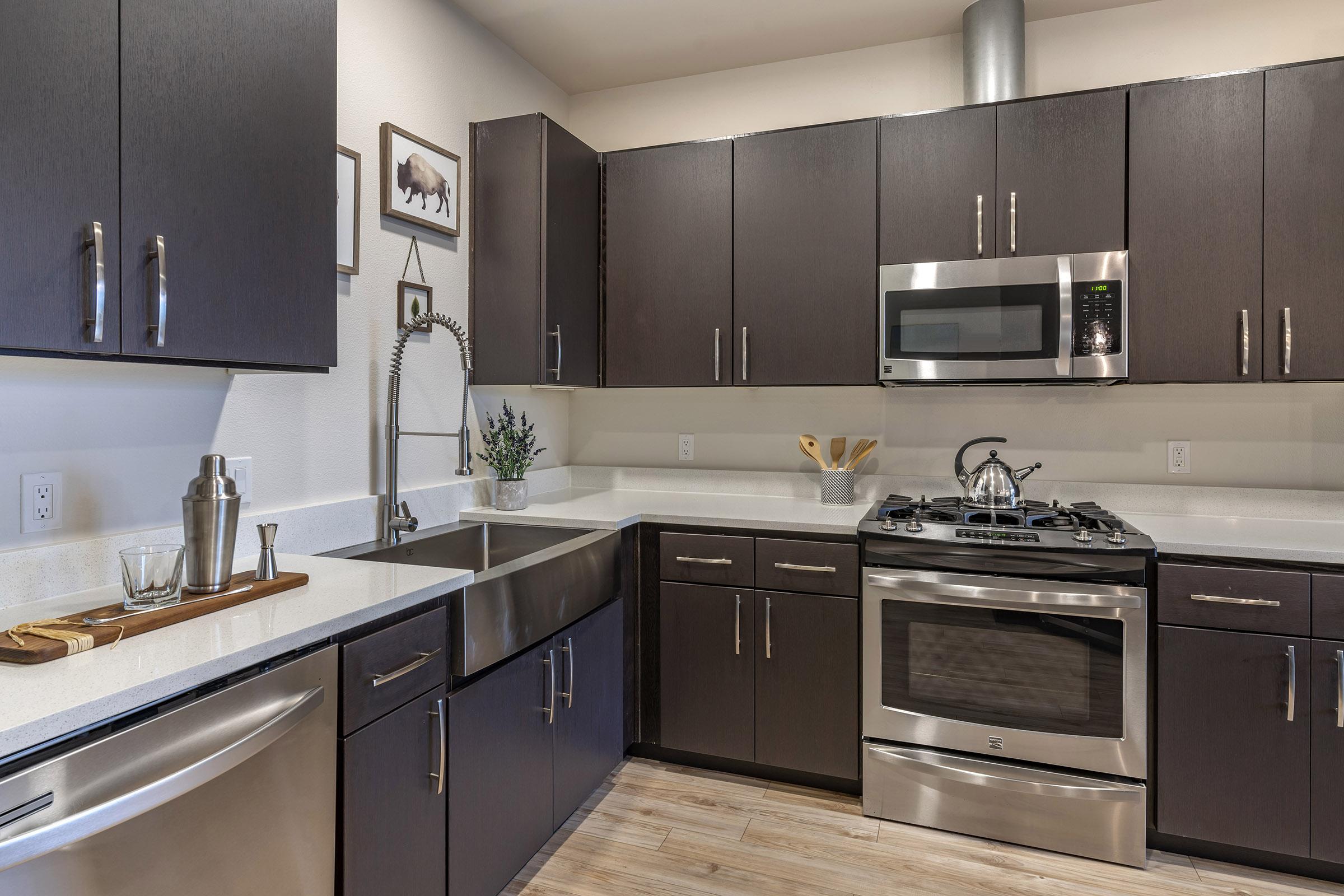 Modern kitchen featuring sleek black cabinetry, stainless steel appliances including a microwave and a gas stove, a double sink with a stylish faucet, and light-colored countertops. Decor includes a few framed pictures and kitchen utensils on display. The flooring is light wood.
