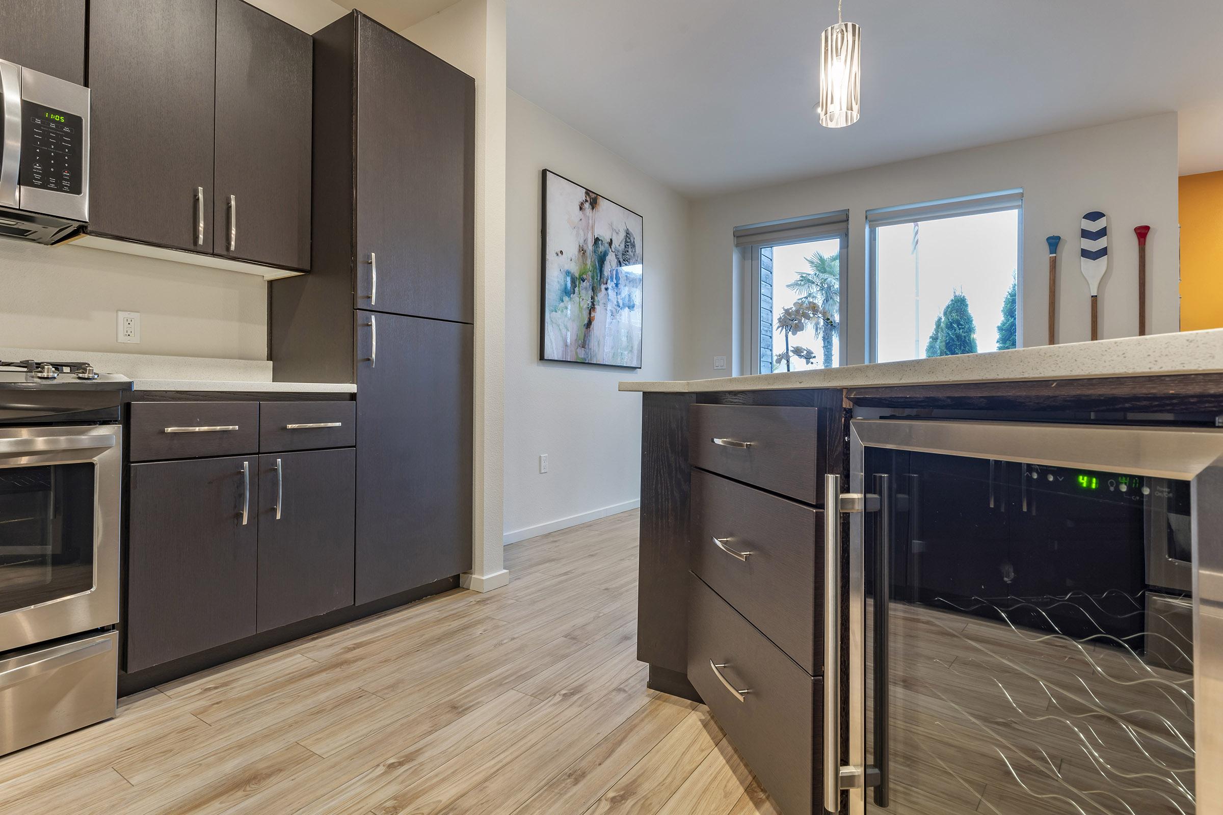 Modern kitchen with dark cabinetry, stainless steel appliances, and a countertop with a wine cooler. Large windows provide natural light, and a piece of abstract art hangs on the wall. The flooring is light-colored wood, creating a contemporary and inviting atmosphere.