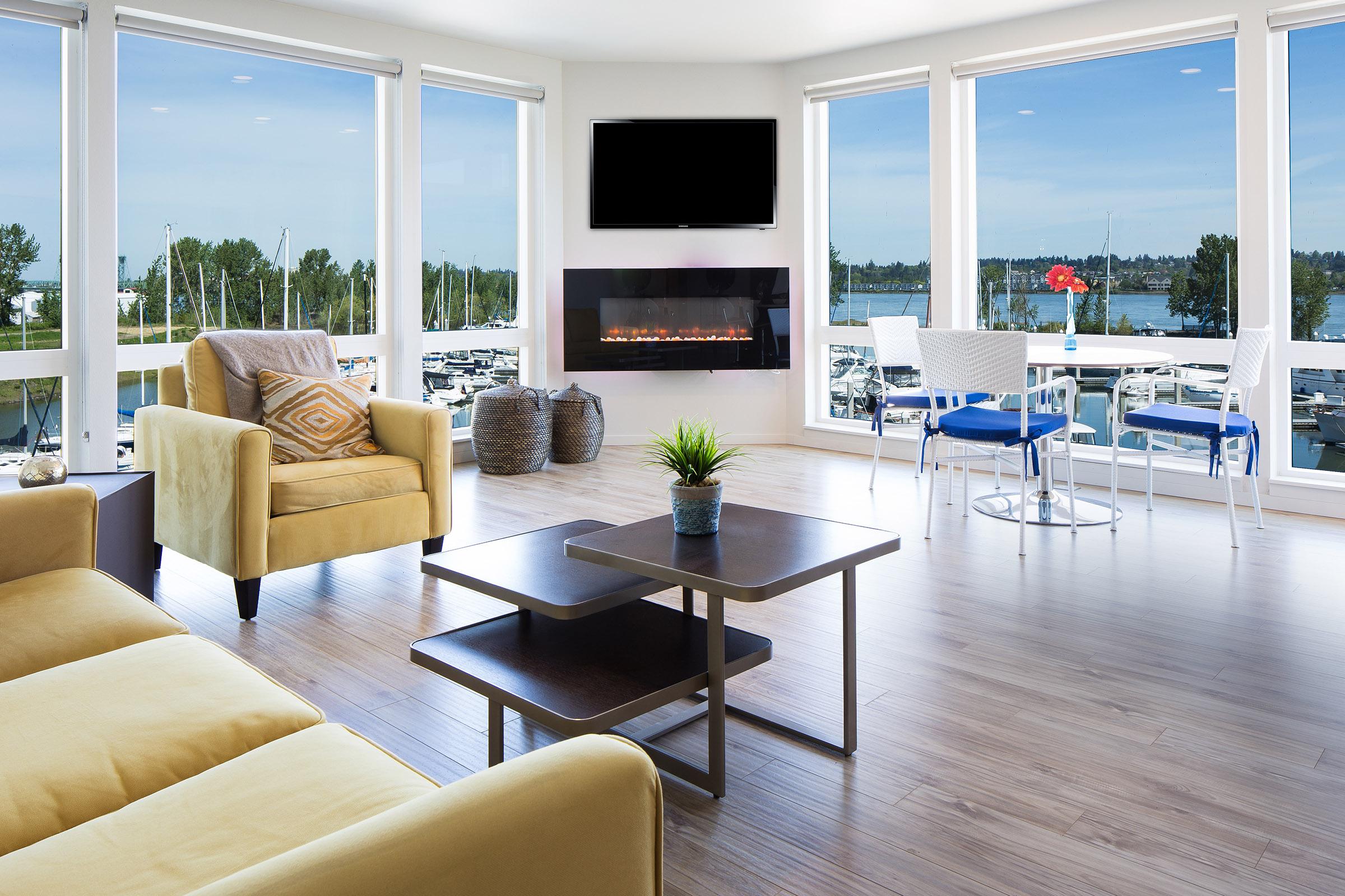 A modern living room with large windows offering a view of a marina. The space features a yellow sofa, a coffee table, and a potted plant. A wall-mounted TV and a fireplace add warmth, while a dining area with two chairs is visible nearby. Natural light floods the room, creating a bright and inviting atmosphere.