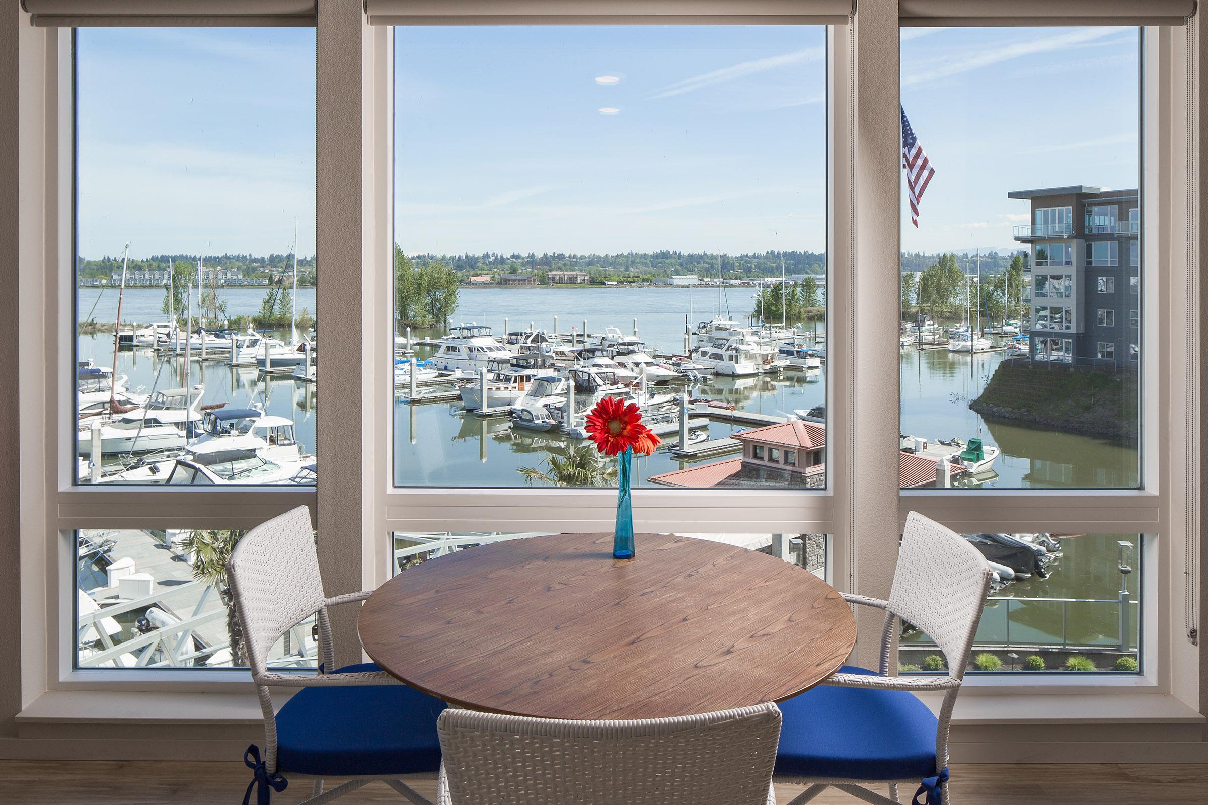 A view from a modern waterfront apartment, featuring a round wooden table with a blue vase containing a red flower. In the background, a marina filled with boats is visible through large windows, with lush greenery lining the shore and a clear blue sky overhead.