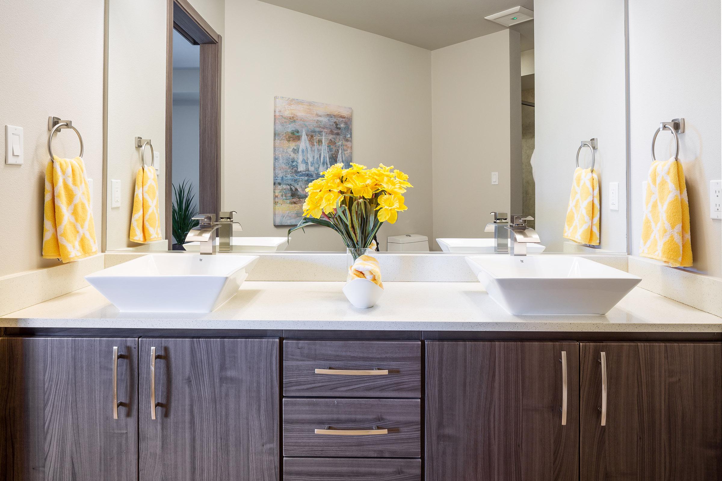 A modern bathroom featuring double sinks with square basins, a stylish countertop, and a decorative arrangement of yellow flowers in a white vase. The decor includes yellow hand towels and mirrors mounted above the sinks, adding a bright and inviting touch to the space.