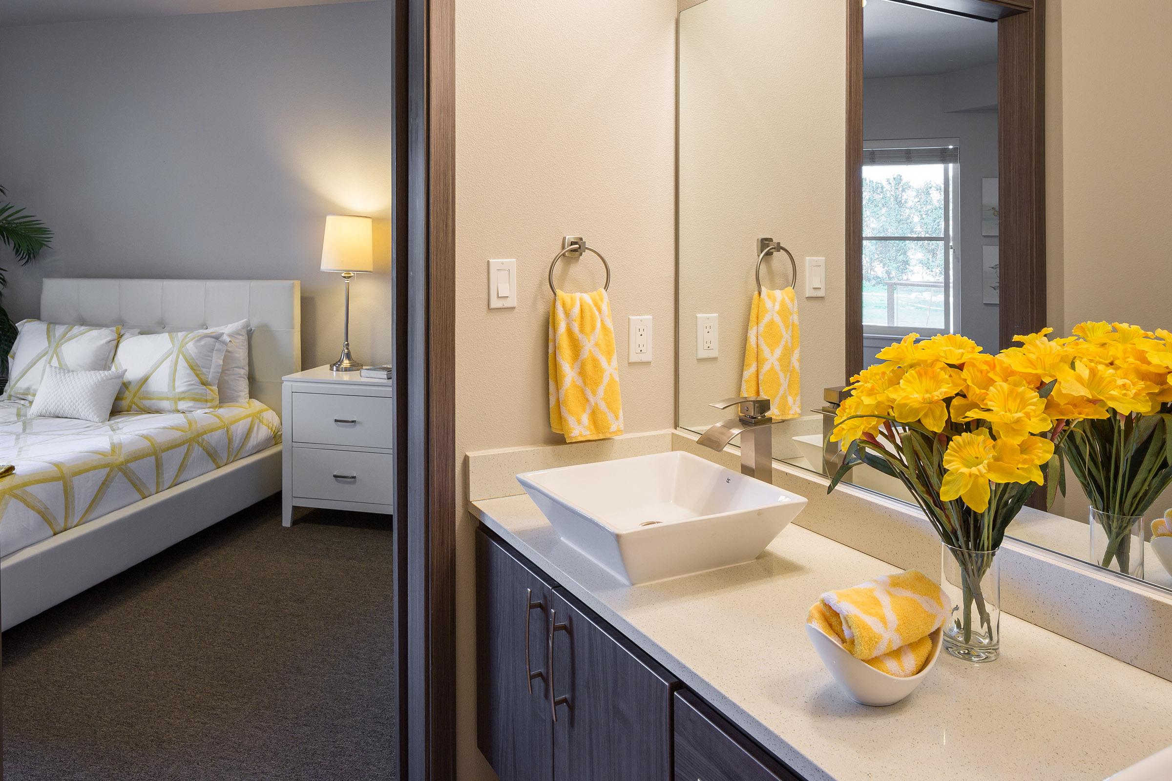 A modern bathroom with a white sink and decorative yellow towels, featuring a vase of yellow flowers. In the background, a cozy bedroom can be seen, complete with a neatly made bed and a bedside lamp. The overall color scheme is fresh and inviting, highlighting contemporary interior design.