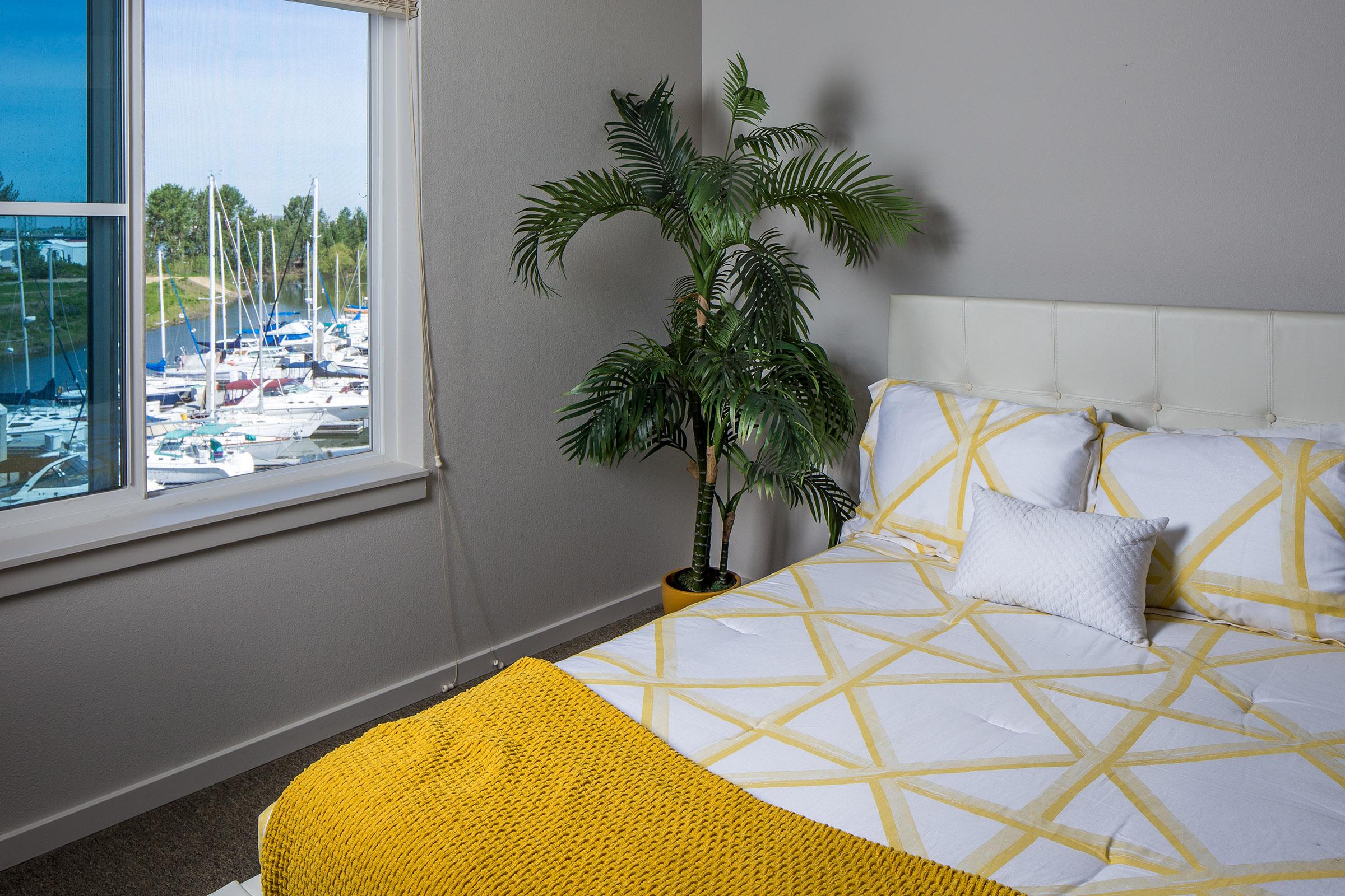 A bright, modern bedroom featuring a white bed with a yellow geometric pattern comforter. There's a white decorative pillow on the bed, a potted palm tree in the corner, and a large window showcasing a view of a marina filled with sailboats and green trees in the background.