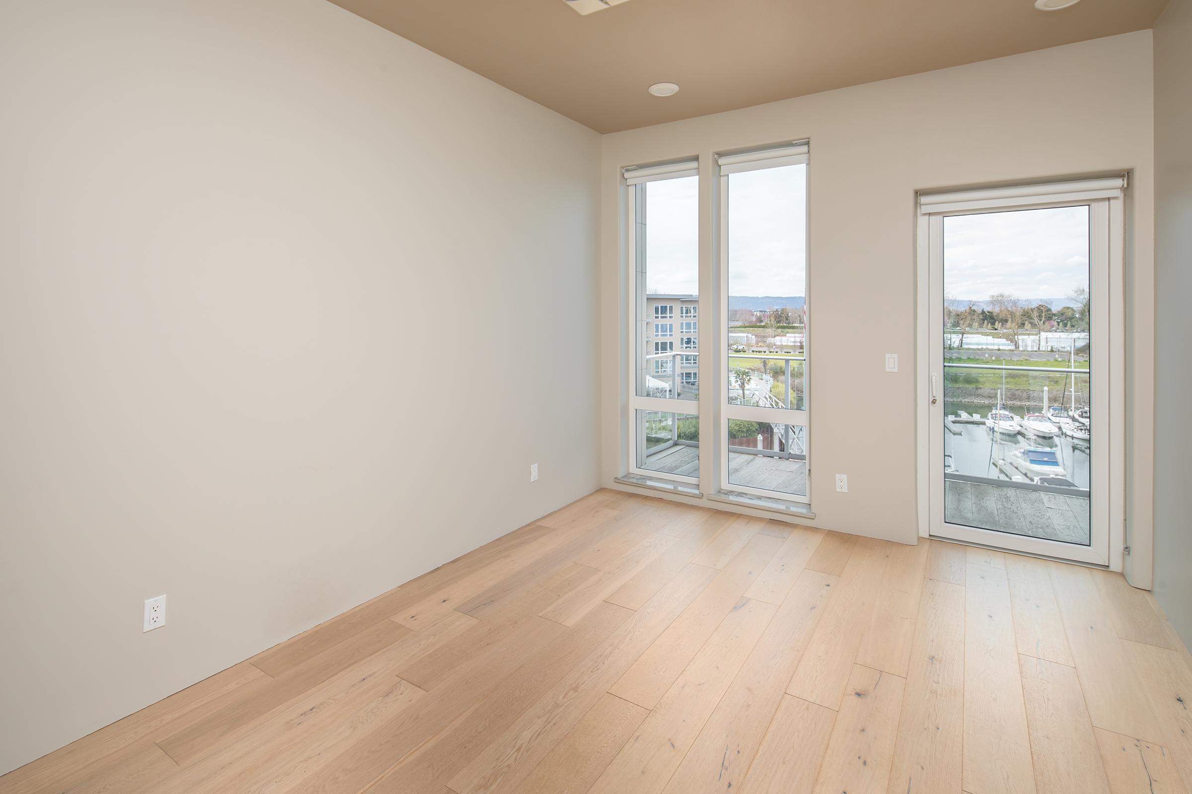 A spacious, empty room with light-colored walls and wooden flooring. Large windows allow natural light to fill the space, showcasing a view of a marina outside. A sliding glass door leads to a small balcony. The room has a minimalist design, offering a blank canvas for decoration.