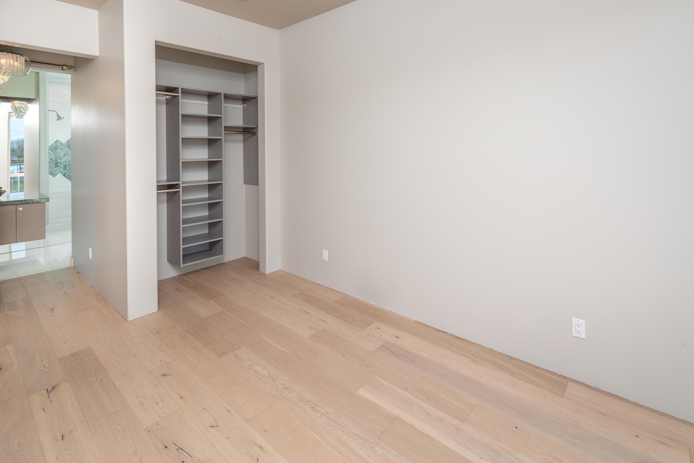 Interior of a room with light-colored wood flooring and neutral walls. To the left, there is a built-in closet with shelving and hanging space. The space is empty, giving it a clean and minimalist look, with a door or opening leading to another area in the background.