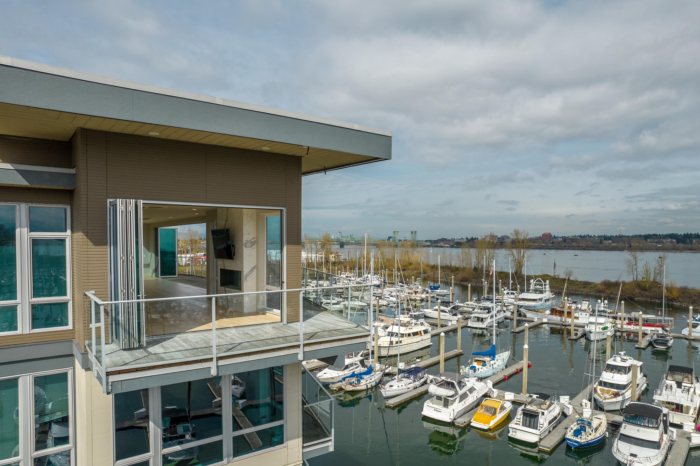 A modern waterfront building with large open balconies overlooking a marina filled with various boats. The scene includes a cloudy sky and a calm body of water, with distant land visible beyond the marina.