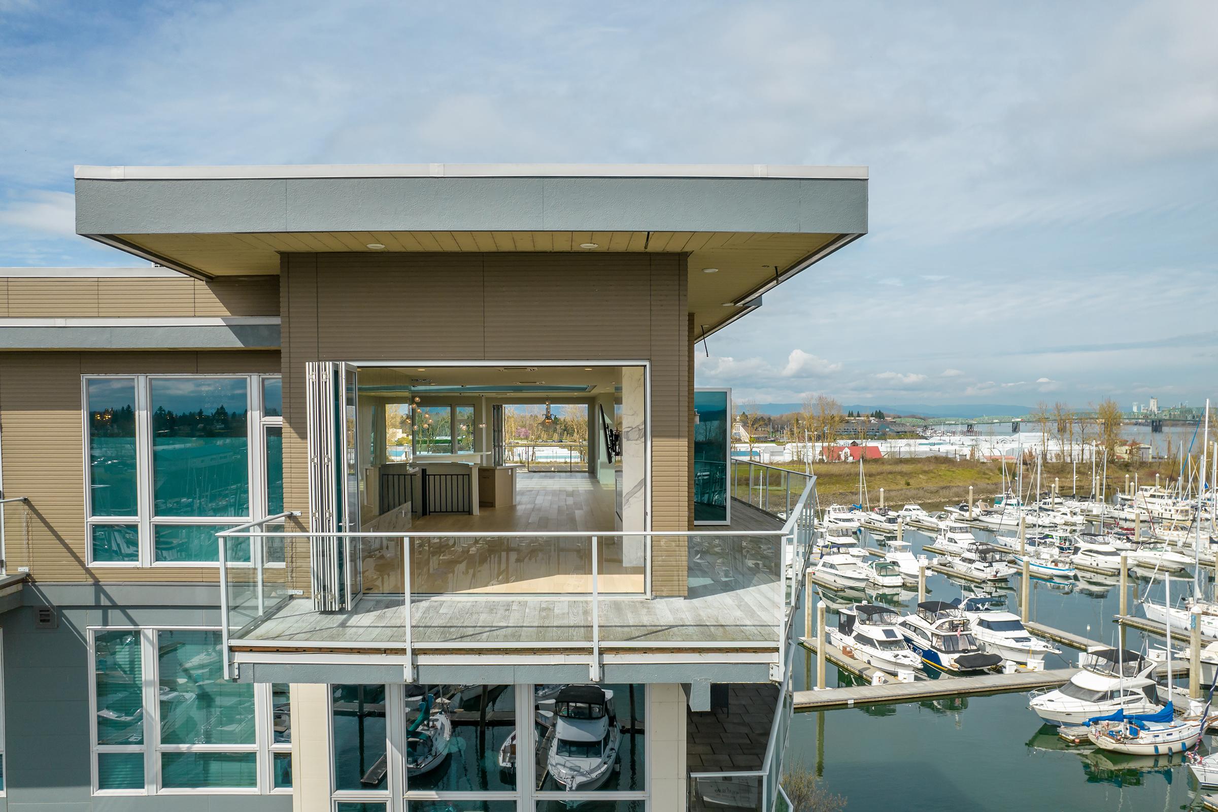 Modern waterfront building with large glass windows and a spacious balcony overlooking a marina filled with boats. The scene features a clear blue sky and calm waters, highlighting the serene coastal environment. Empty interior space visible through the open balcony doors.