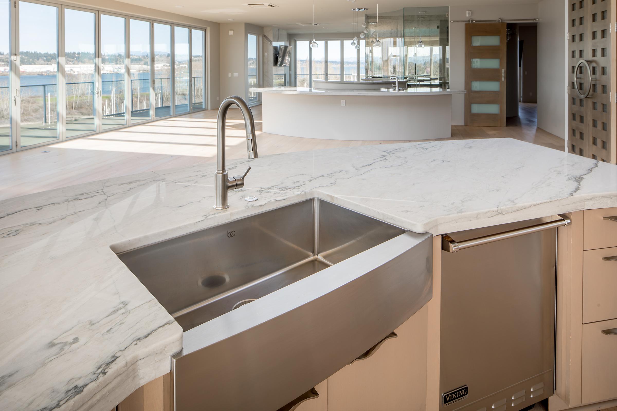 A modern kitchen with a sleek marble countertop and stainless steel sink. The background features large glass windows overlooking a waterfront view, revealing natural light in a spacious, open-plan living area. The design is contemporary with neutral tones and minimalist cabinetry.