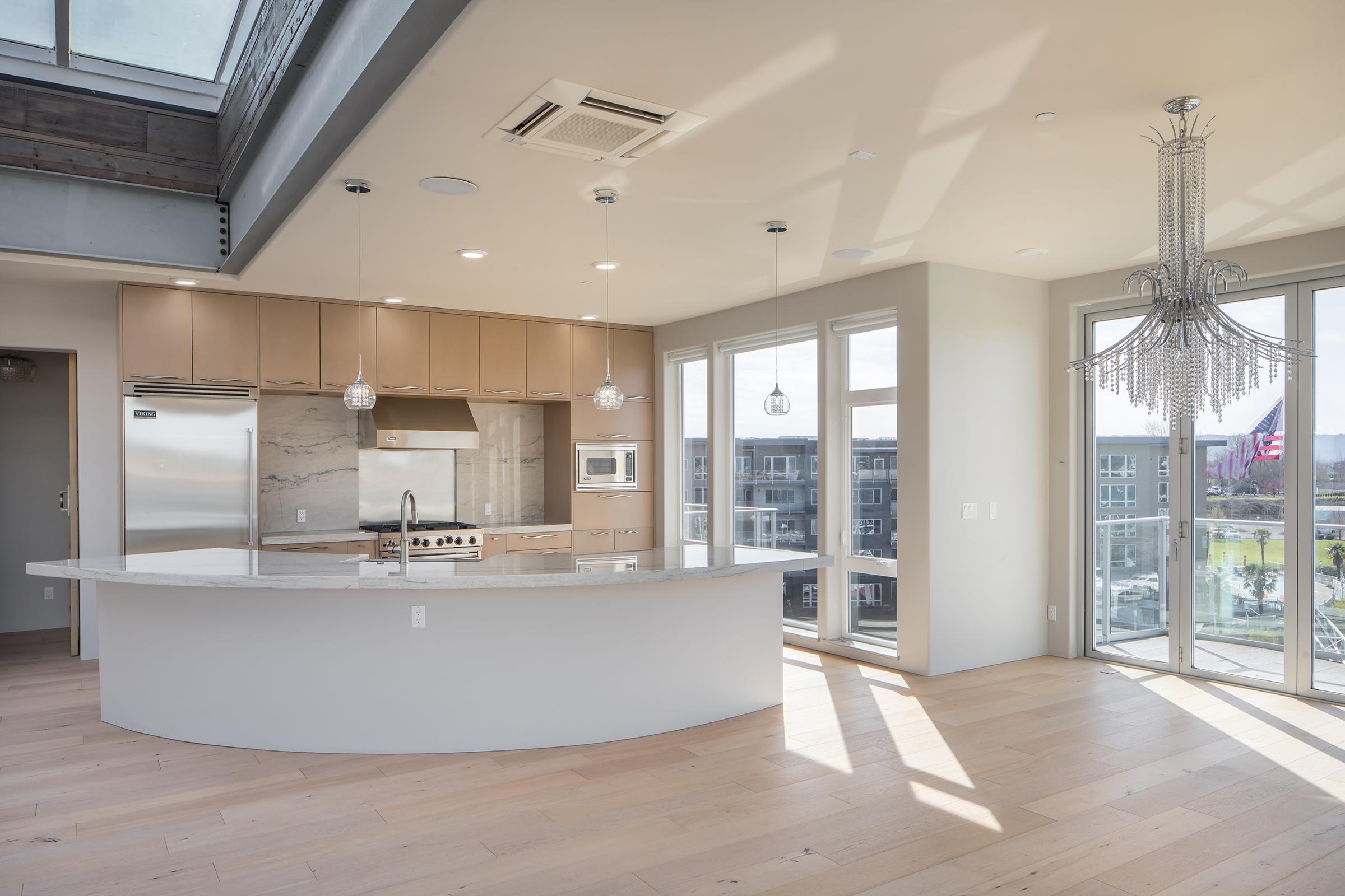 Modern kitchen with an open layout featuring a large curved island counter, stylish cabinetry, and stainless steel appliances. Abundant natural light flows in through large windows, highlighting a contemporary chandelier above. The space has hardwood flooring and a minimalist design, creating an airy atmosphere.