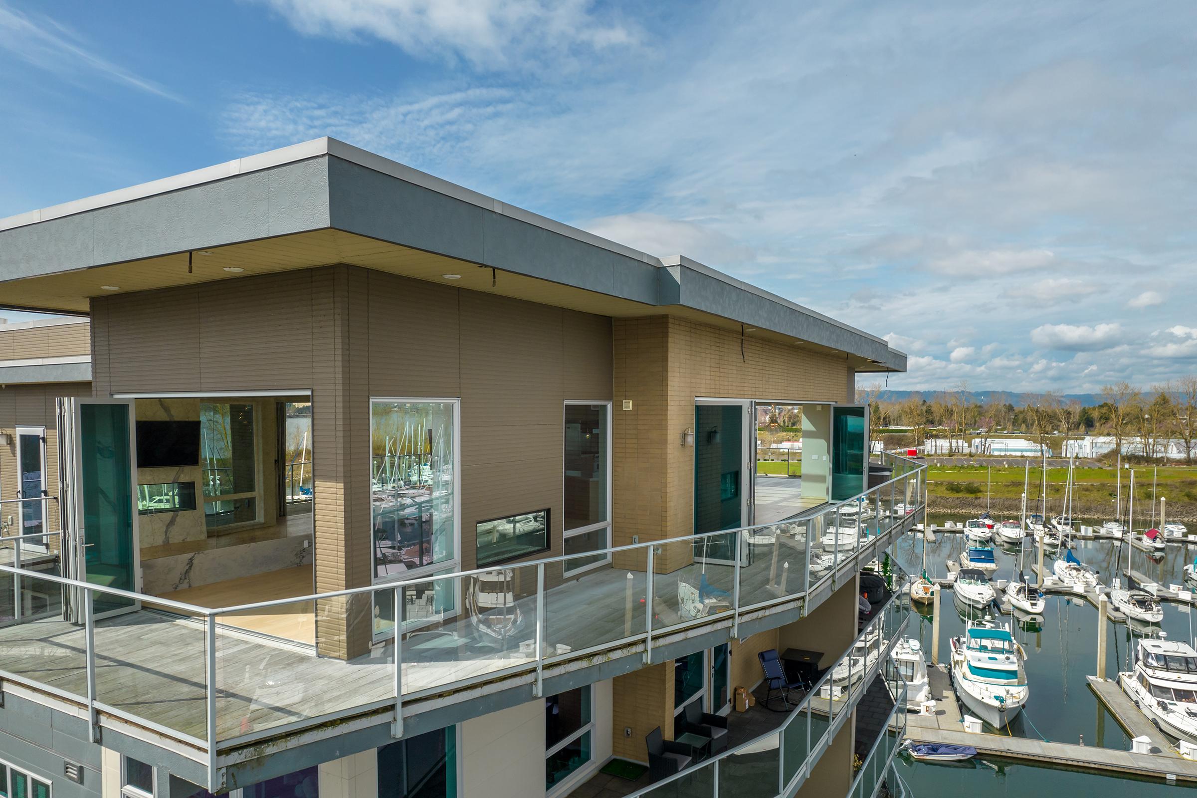Modern waterfront property featuring a spacious balcony overlooking a marina filled with boats. The building has large glass windows, providing abundant natural light, and the surrounding area includes lush greenery and blue skies with scattered clouds.