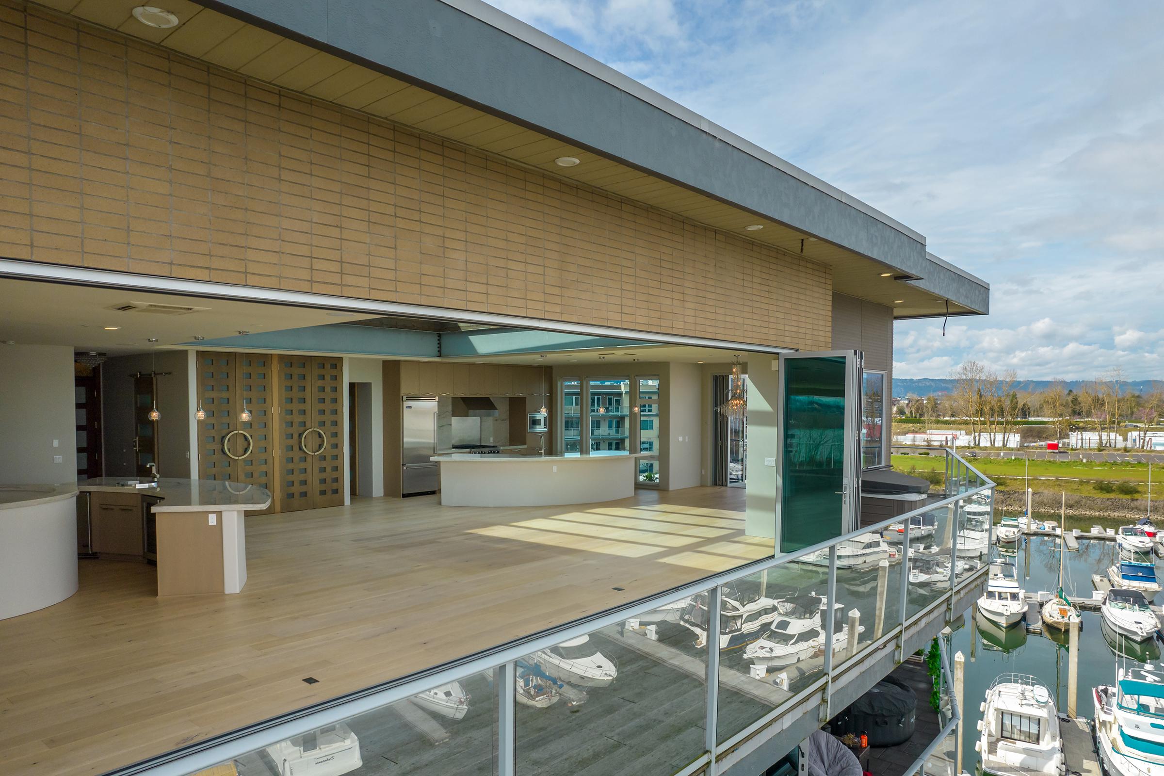 A modern, open-concept kitchen and living area with large windows overlooking a marina filled with boats. The room features wooden flooring, sleek countertops, and contemporary fixtures. The outdoor balcony offers a panoramic view of the water and the surrounding landscape.