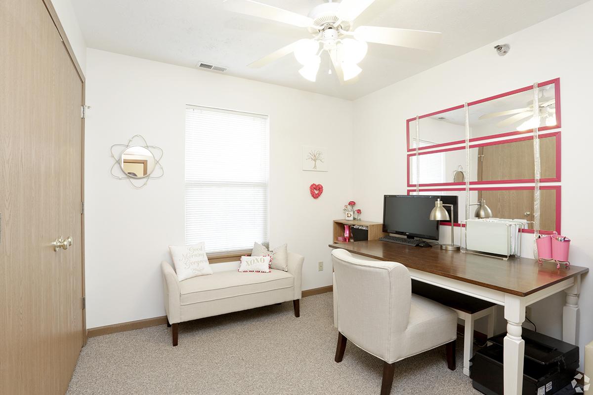 A bright and cozy home office space featuring a wooden desk with two stylish desk lamps, a computer monitor, and decorative items. A small beige armchair sits nearby, accompanied by decorative pillows. The walls are adorned with light-colored accents, including a mirror and framed art, and a window lets in natural light.