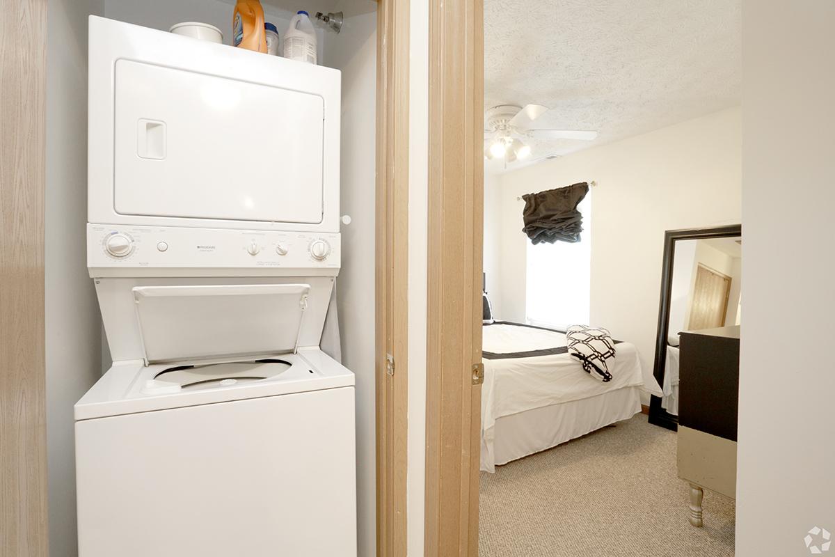 A compact laundry area with a stacked washer and dryer, located in a small alcove. Nearby, a doorway leads to a softly lit bedroom featuring a bed, a mirror, and a window with a draped curtain, creating a cozy atmosphere.