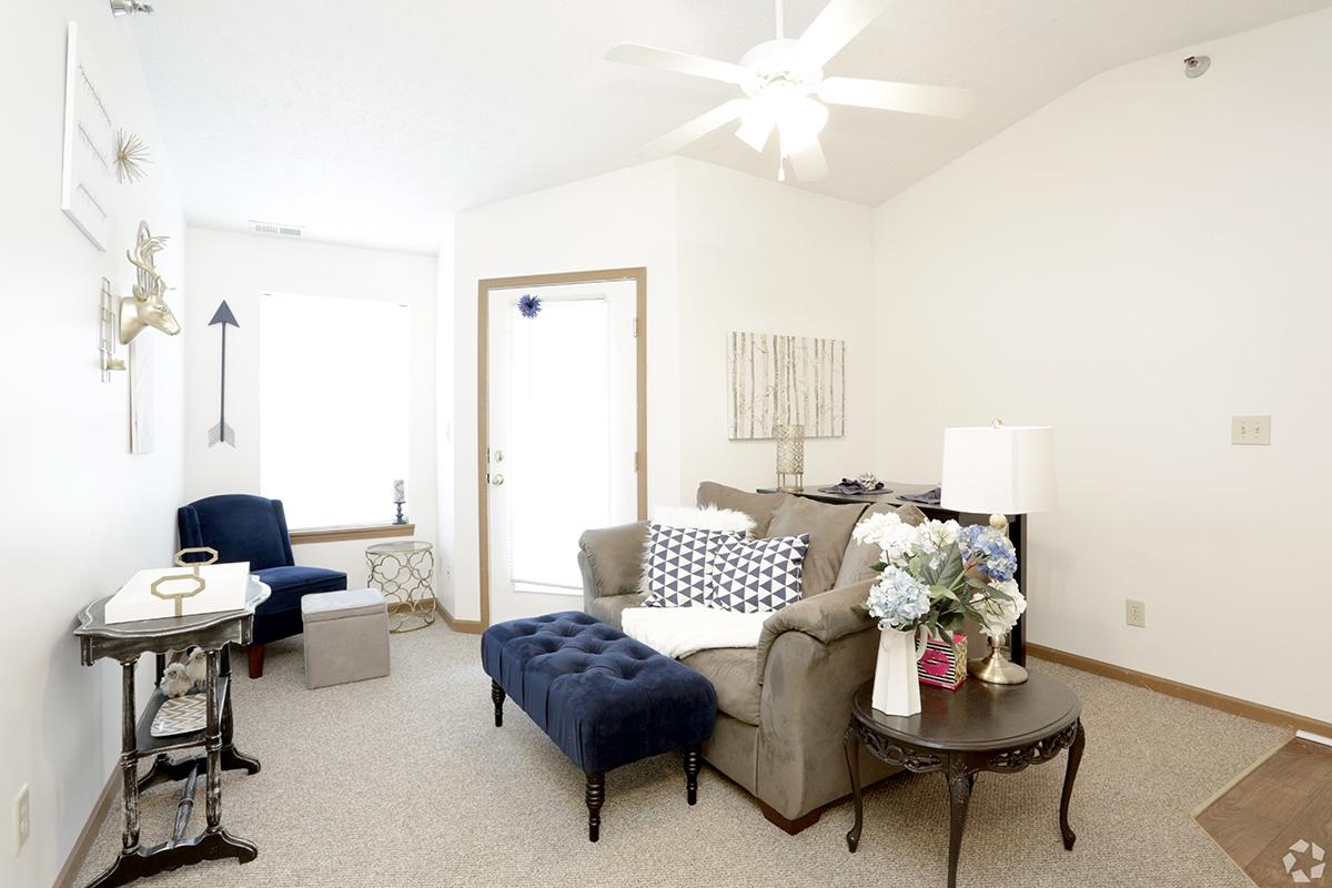A cozy living room featuring a blue upholstered chaise lounge, a brown sofa, and a small side table. The room has light-colored walls, a ceiling fan, and a window with natural light. Decorative items include a lamp and a flower arrangement on a round table, creating a welcoming atmosphere.