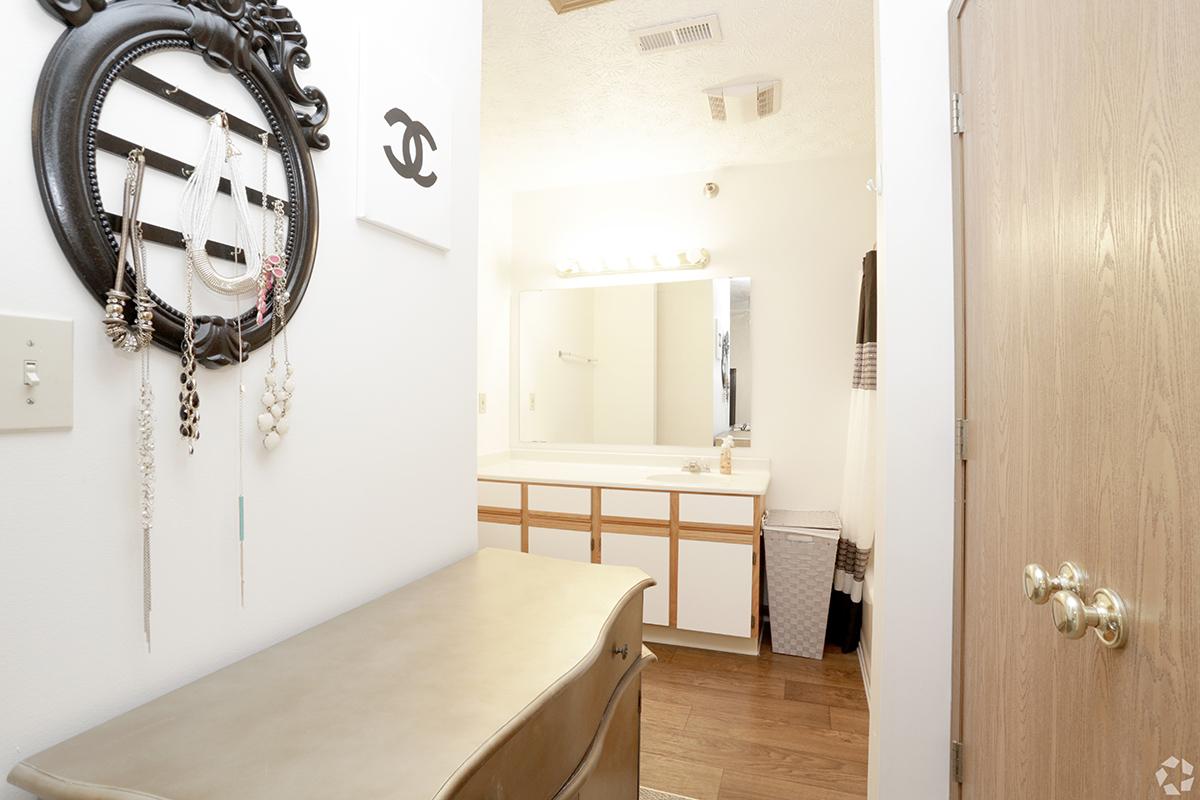 A well-lit bathroom entrance featuring a decorative wall mirror with jewelry hanging from it. The area includes a long countertop with ample space and cabinetry below. A wall-mounted light fixture is above the mirror, and a laundry basket is visible in the corner. The door leads to the bathroom space, with neutral-colored decor.