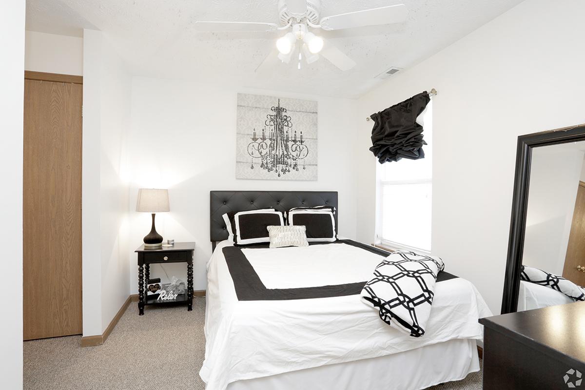 A cozy bedroom featuring a black and white color scheme. The room includes a queen-sized bed with decorative pillows, a nightstand with a lamp, a tall mirror, and a large window with a stylish curtain. A piece of art with a chandelier design hangs above the bed. The flooring is carpeted.