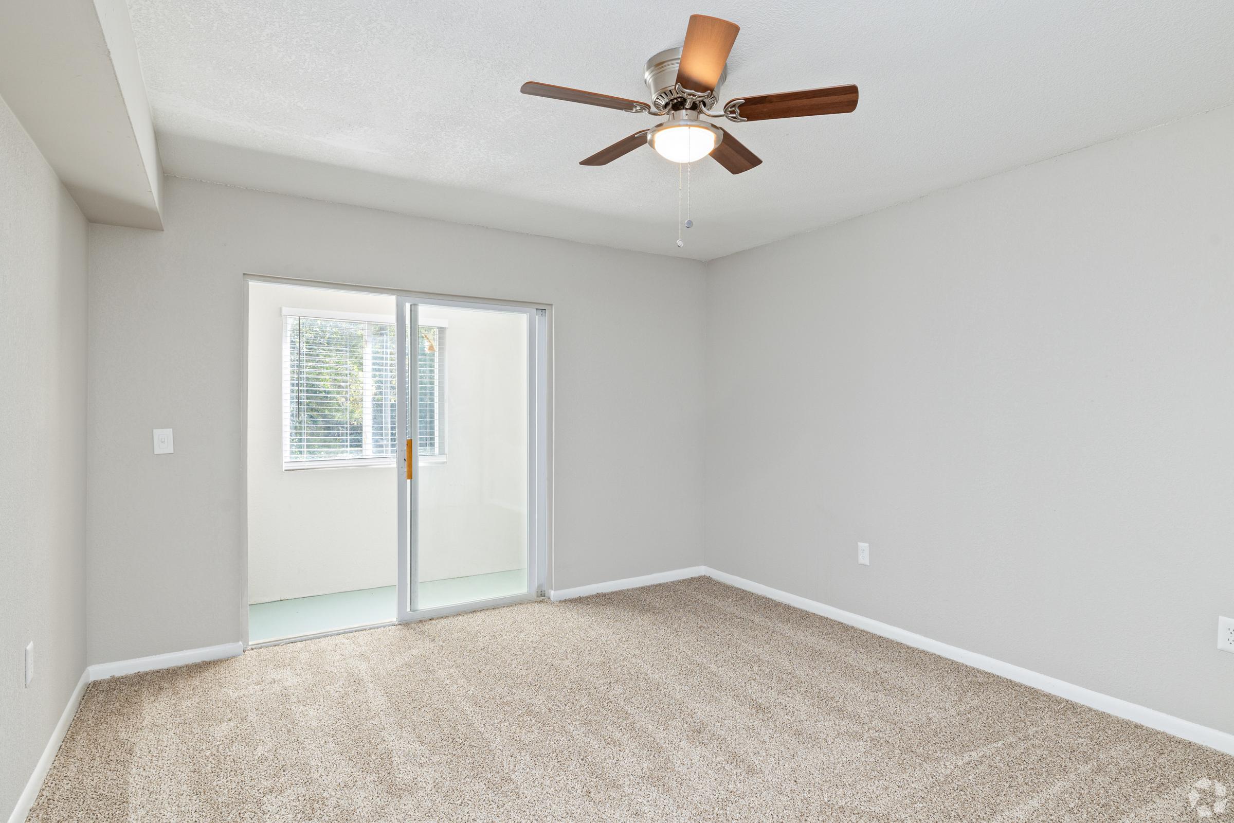 Spacious room with light-colored walls and plush beige carpet. A ceiling fan is installed, and there is a sliding glass door leading to a small patio or balcony area with a window nearby, allowing natural light to enter the room.