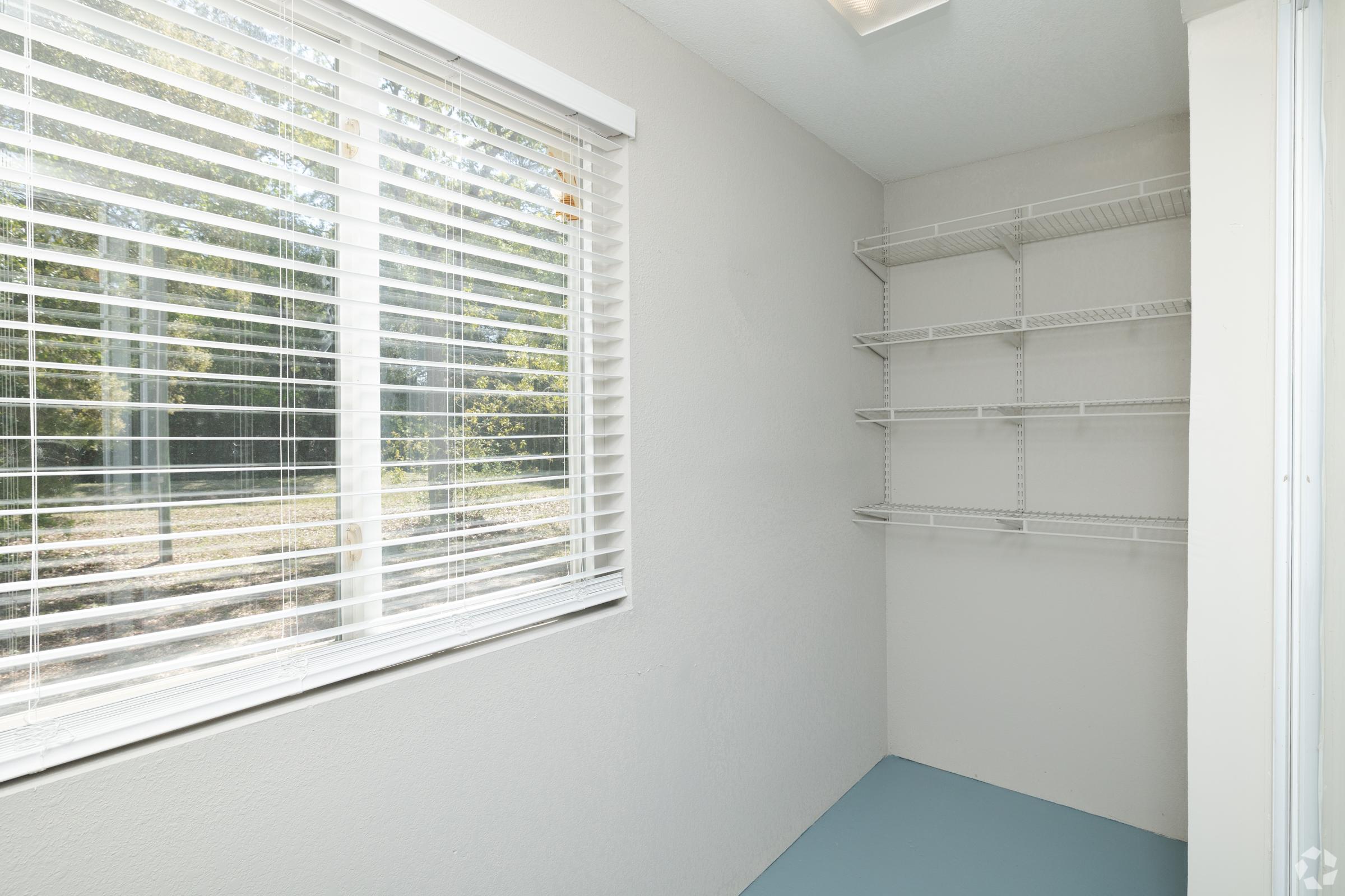 A bright, small room with a large window featuring white blinds that allow natural light in. The walls are painted light gray, and there are several wire shelves on one side. The floor is a solid light blue color, creating a clean and organized space suitable for storage or a small utility area.