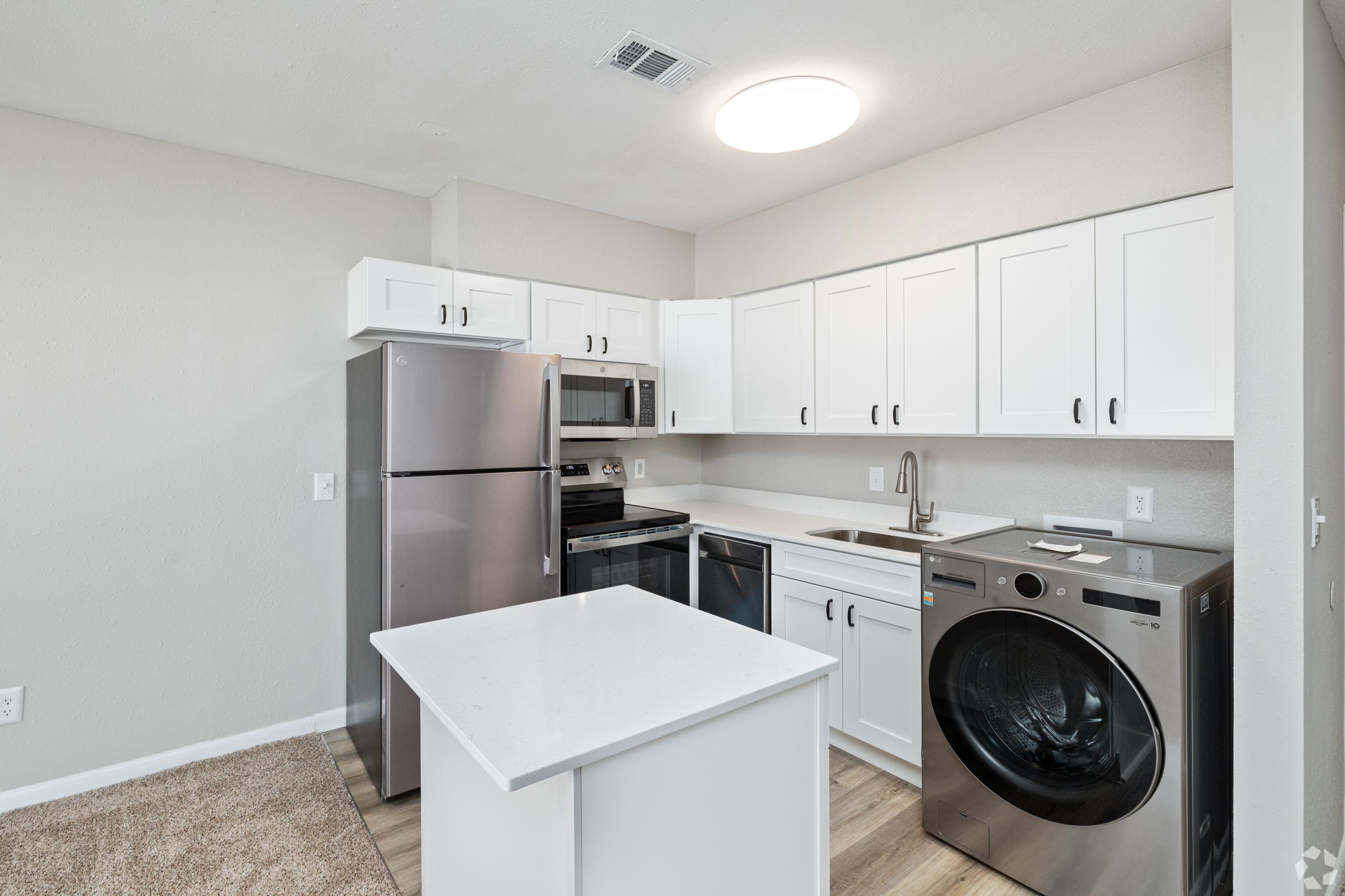 A modern kitchen featuring white cabinets, stainless steel appliances including a refrigerator, microwave, and oven. There is a dishwasher and a washer-dryer combo. The countertop is light-colored, and the floor is covered with a neutral-colored carpet. Bright overhead lighting illuminates the space.