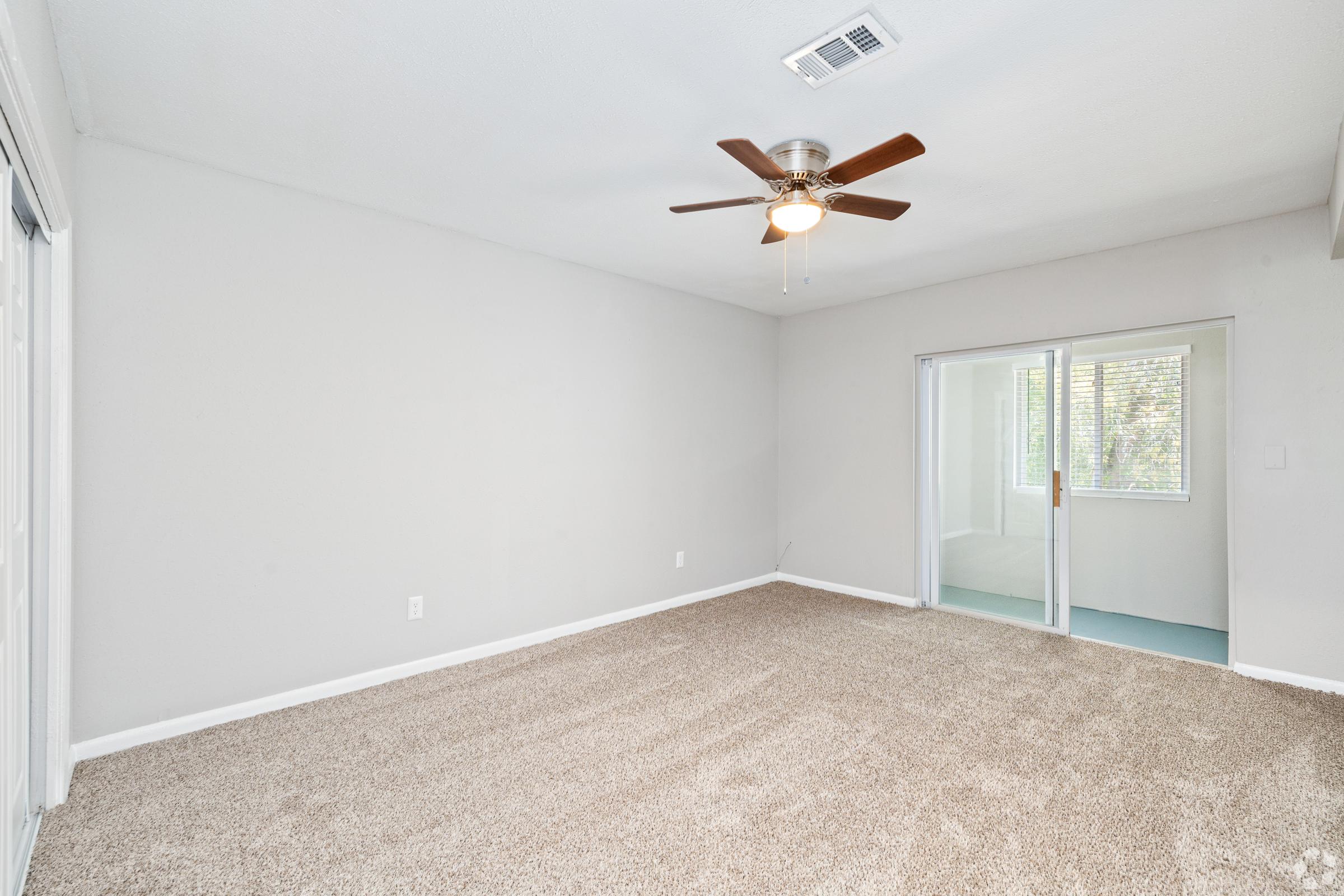 A spacious, empty room with light beige carpet and neutral-colored walls. The room features a ceiling fan with wooden blades and a large window with sliding glass doors that lead to another room. Natural light fills the space, creating a bright and airy atmosphere.