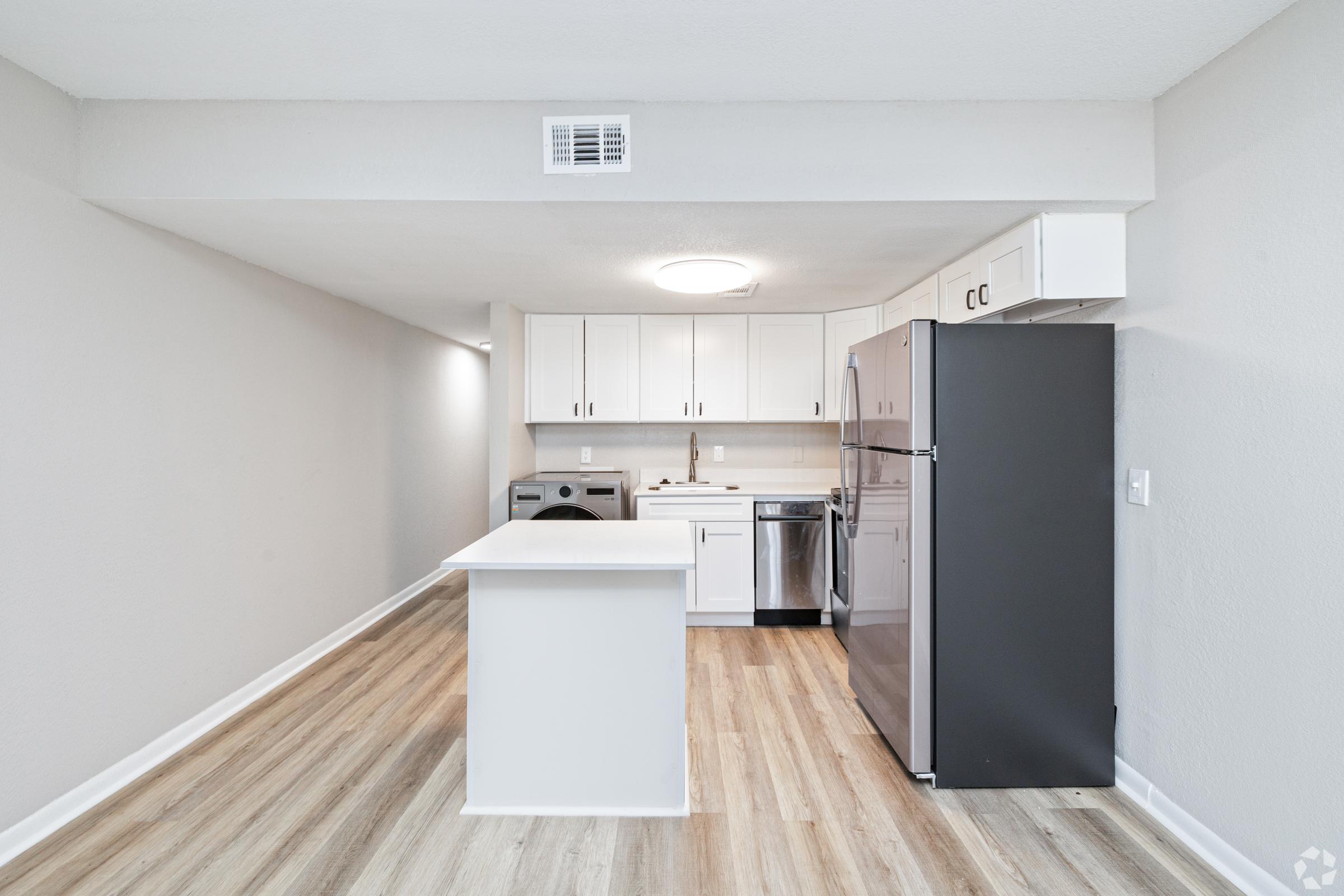 Modern kitchen and laundry area with white cabinetry, stainless steel appliances, and a central island. The space features light-colored wood flooring and neutral wall colors, creating a bright and open atmosphere.