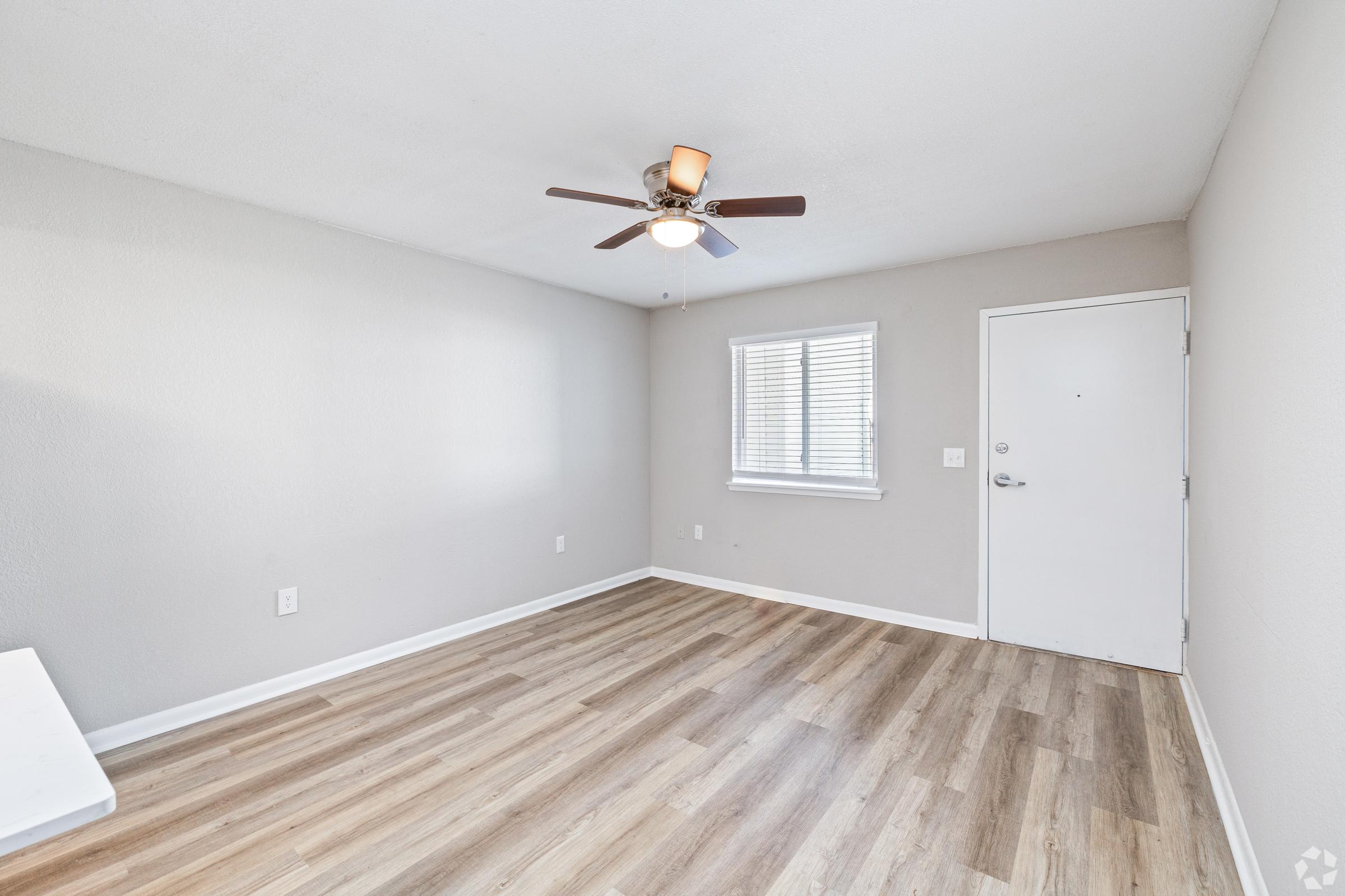 A well-lit, empty room with light brown wooden flooring, a ceiling fan with a light fixture, and a window with blinds. The walls are painted in a soft gray color, and there is a white door leading outside. The space appears clean and ready for furniture or decoration.
