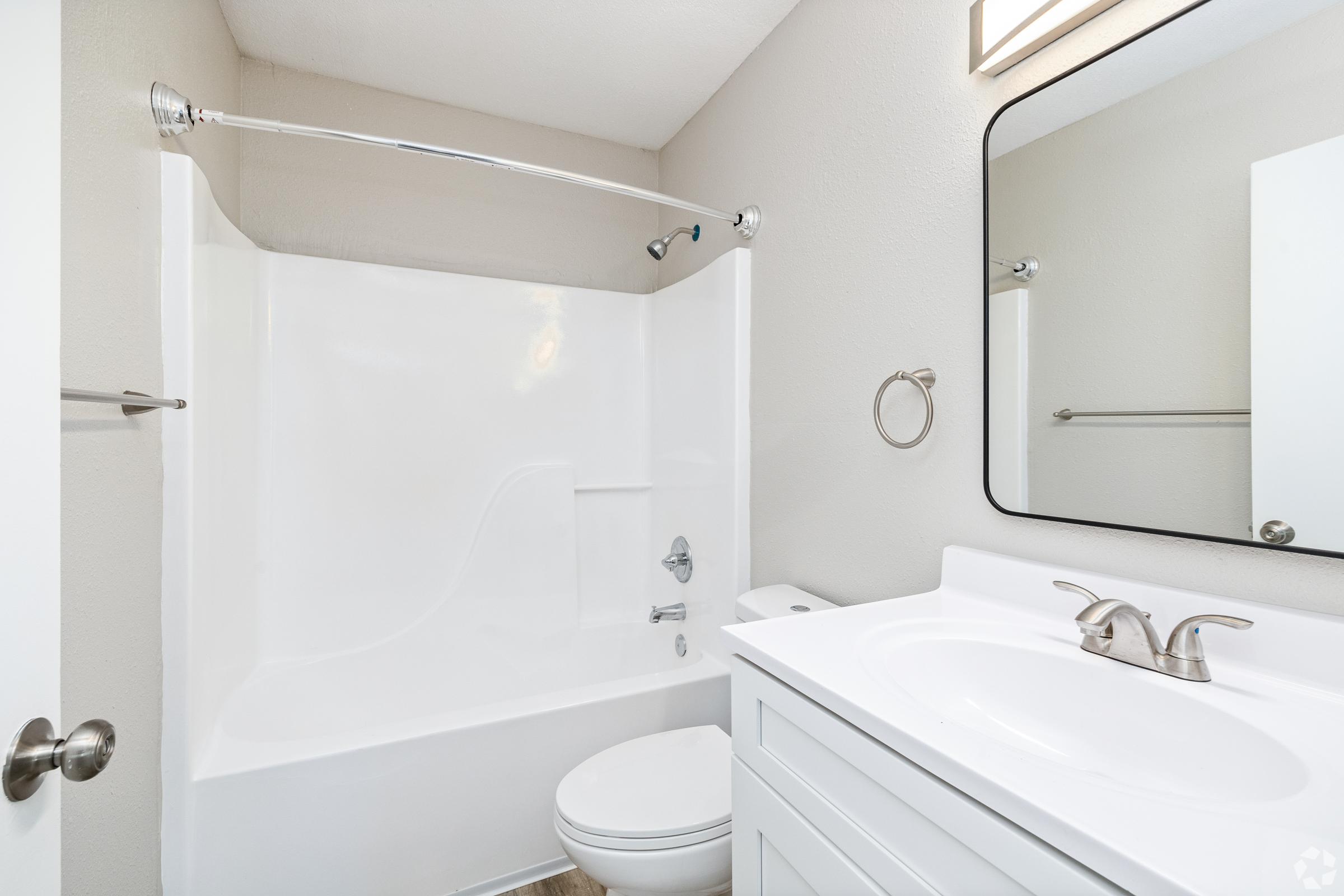 Modern bathroom featuring a clean white shower/tub combo, a compact toilet, and a sleek vanity with a sink. The walls are painted in a light, neutral color, and there's a mirror with a simple frame above the sink. A towel bar is mounted on the wall, and natural light brightens the space.