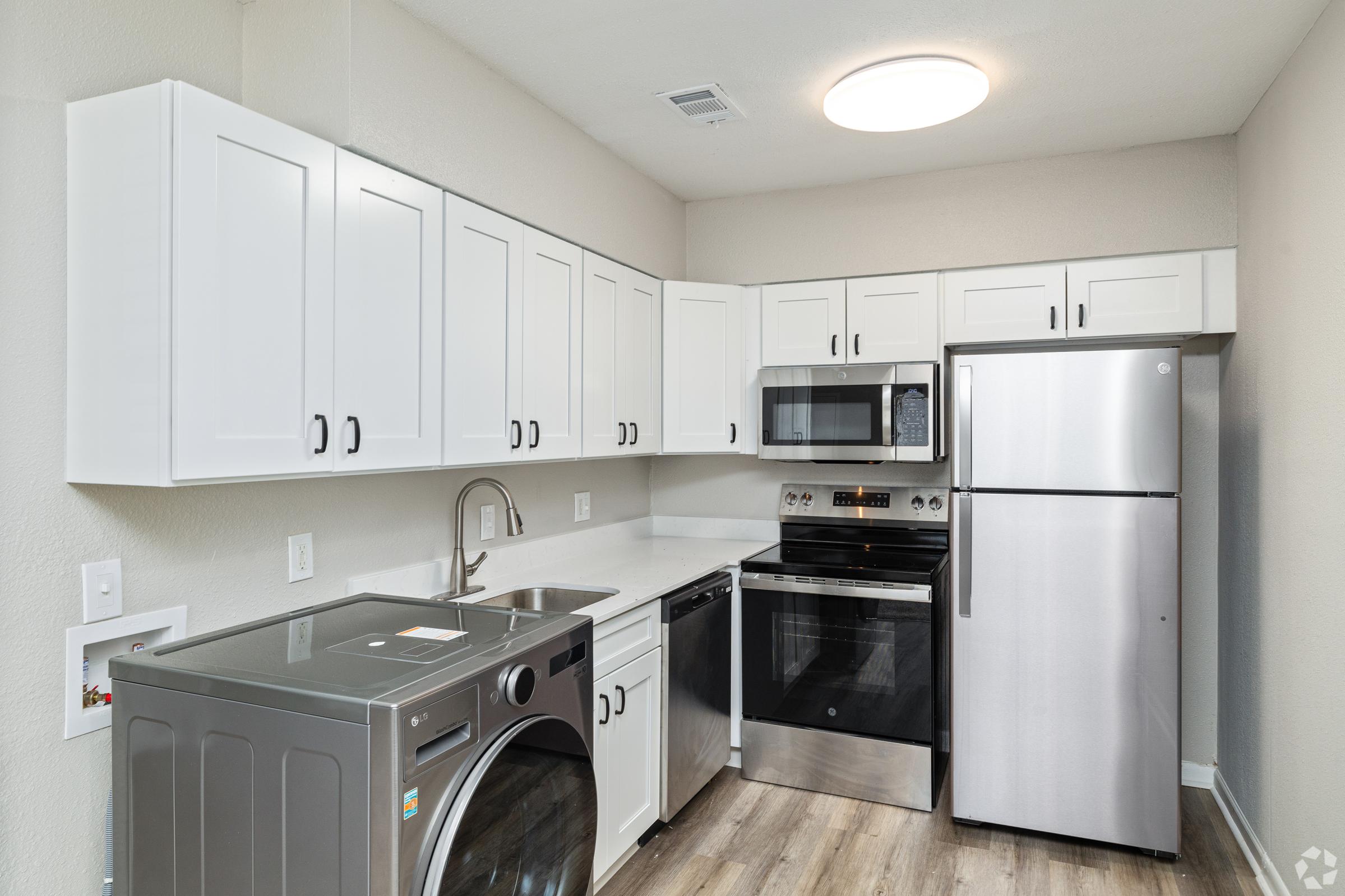 A modern kitchen featuring white cabinets, stainless steel appliances including a microwave, oven, and refrigerator, along with a washer and dryer unit. The space has a light-colored wall and a bright ceiling light, complemented by wooden flooring.