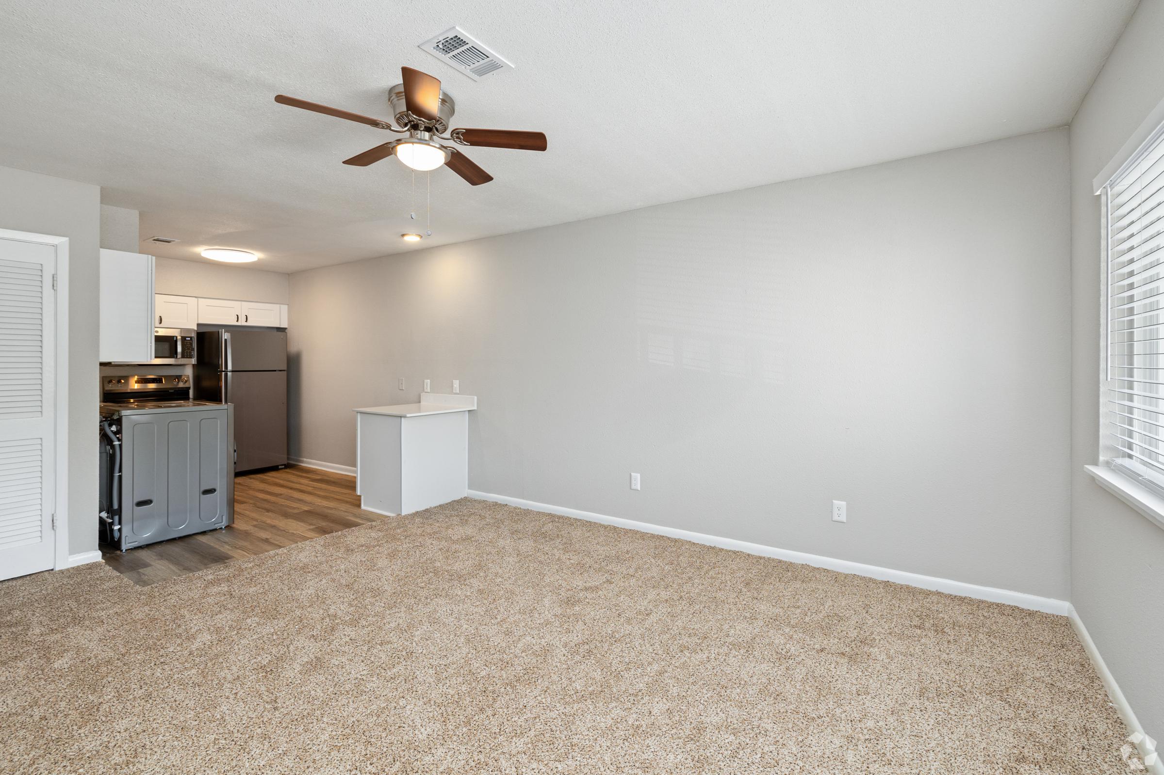 Interior of a spacious living area featuring tan carpet flooring, a ceiling fan, and neutral-colored walls. To the left, a small kitchen with modern appliances, including a refrigerator and stove. A window allows natural light to brighten the room, enhancing the open feel of the space.