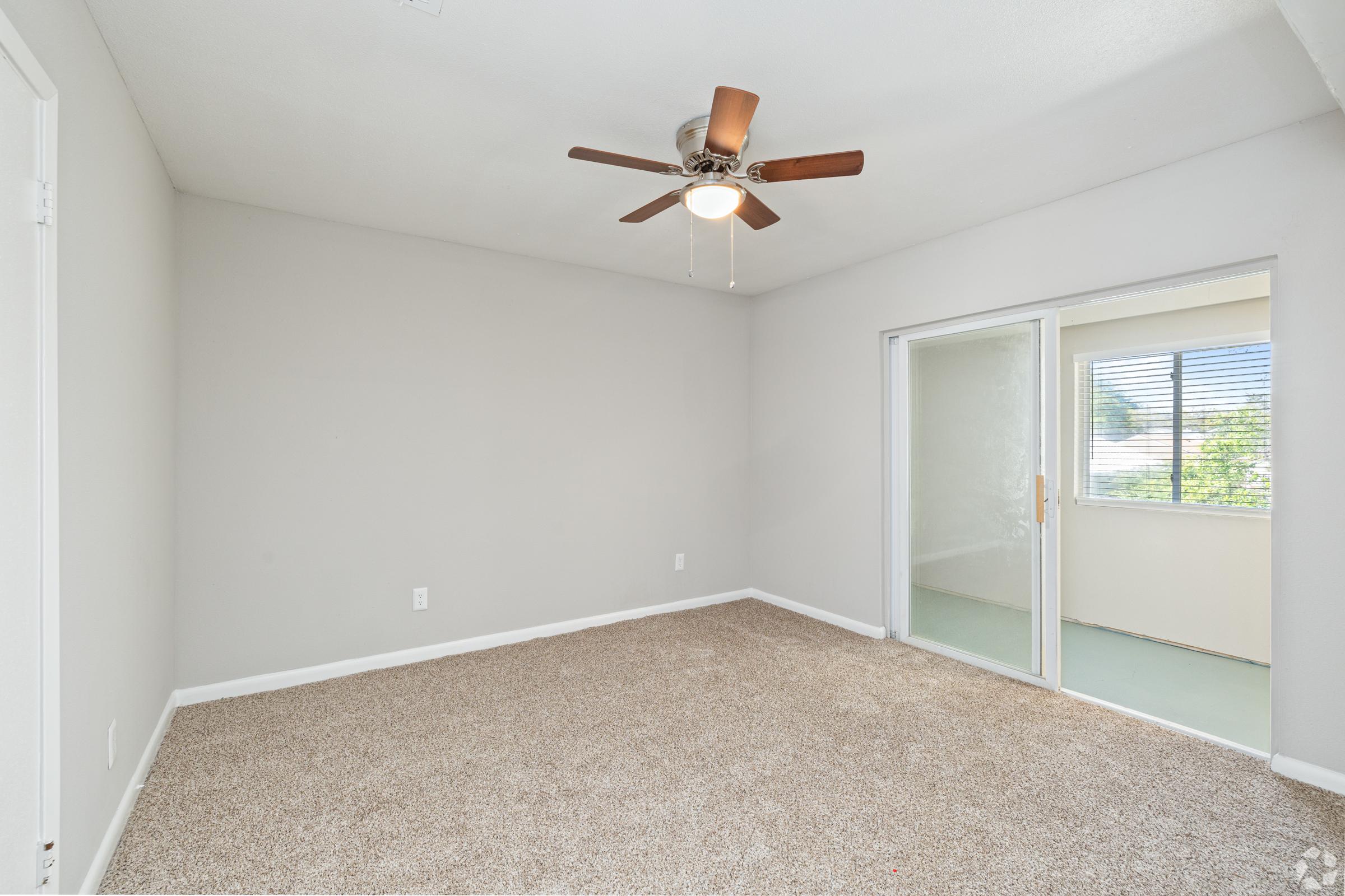 A spacious, empty room with light beige carpet, a ceiling fan with wooden blades, and a sliding glass door leading to a bright area with windows. The walls are painted in a soft gray color, providing a neutral backdrop for potential furnishings. Natural light filters into the room from the adjacent space.