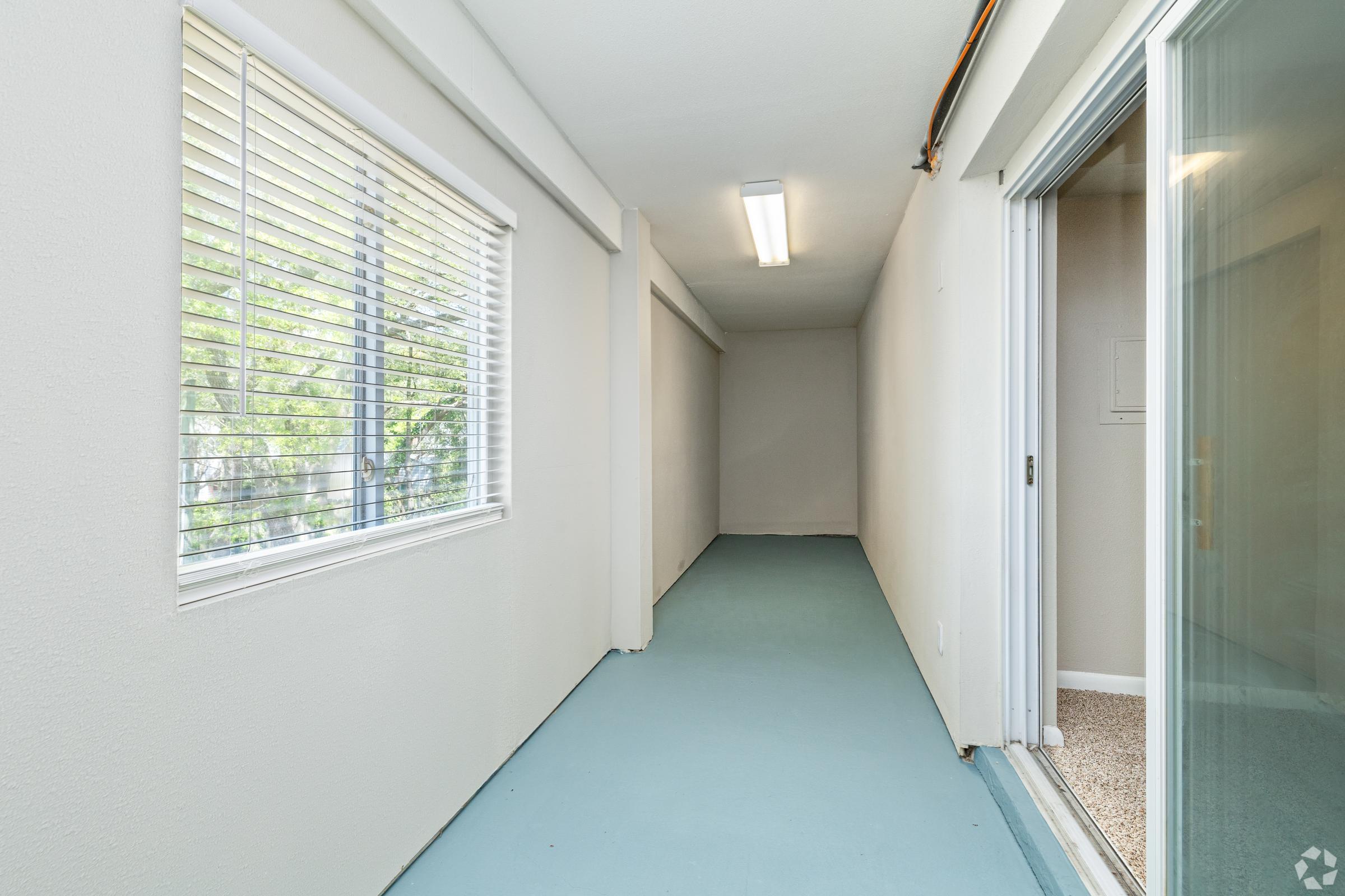 A narrow hallway with light blue flooring, an open sliding glass door on the right leading to an adjacent room, and a large window on the left allowing natural light to enter. The walls are painted light-colored, creating a bright and spacious feel in the corridor.