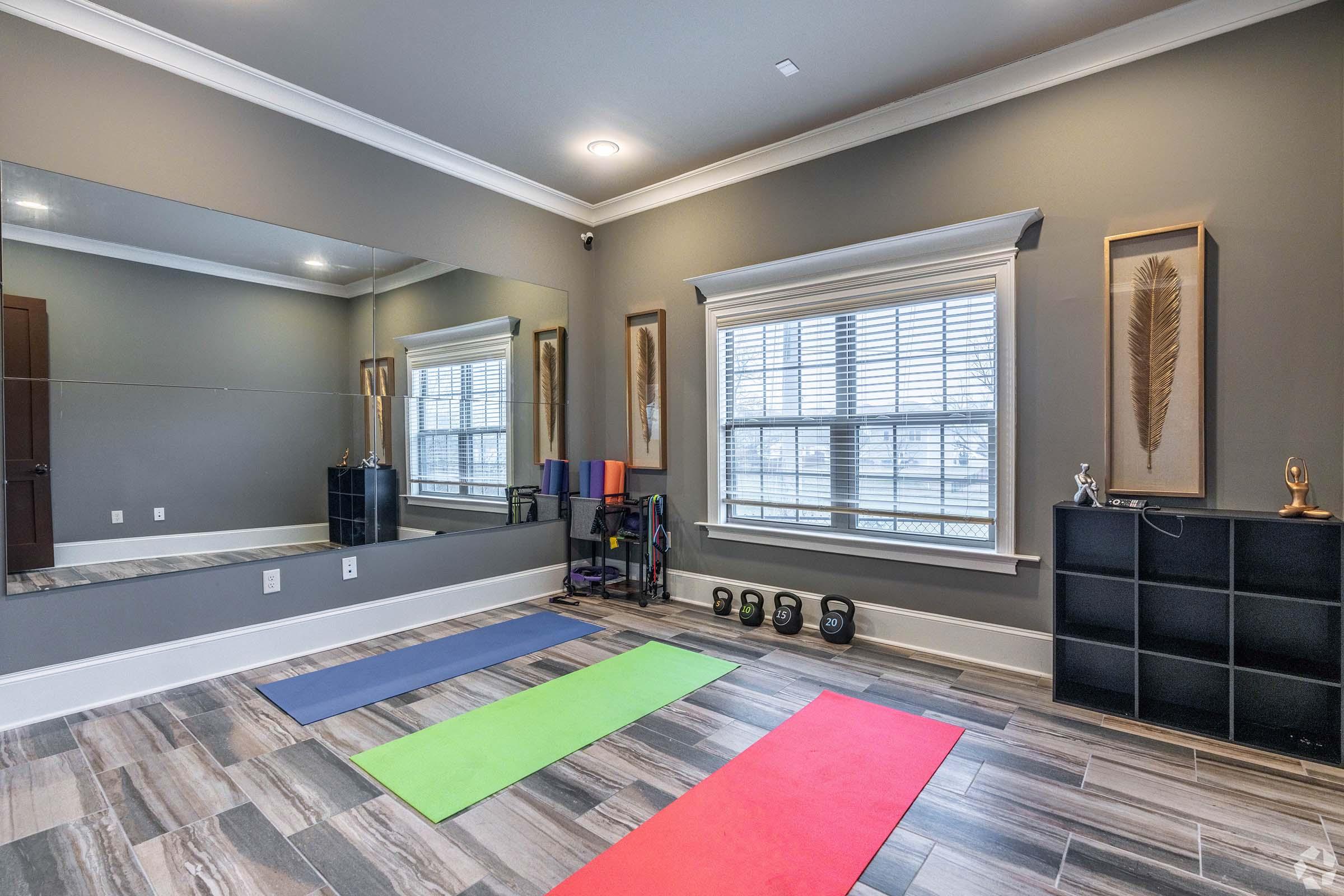 A modern fitness room featuring three exercise mats in red, green, and blue arranged on a wooden floor. A large mirror reflects the space, which includes a window with blinds. There are dumbbells on a rack and decorative wall art framed in the background, creating an inviting workout environment.