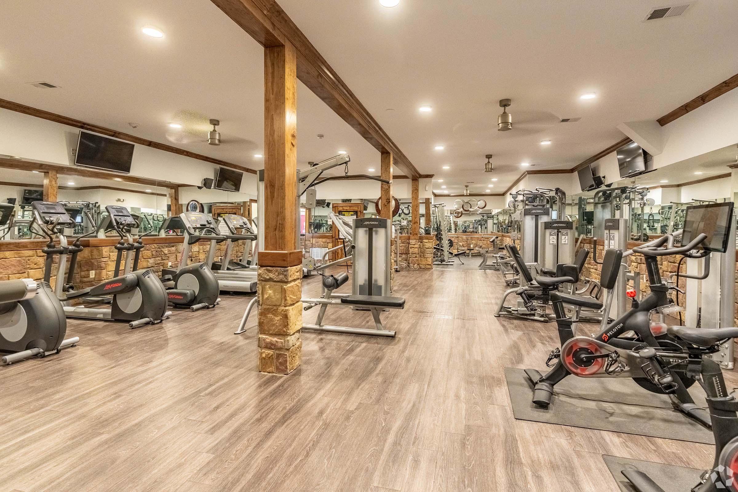A spacious gym featuring a variety of exercise equipment, including treadmills, stationary bikes, and weight machines. The interior has wooden beams and stone accents, with large mirrors reflecting the setup and multiple television screens mounted on the walls. The floor is hardwood, adding to the modern design.