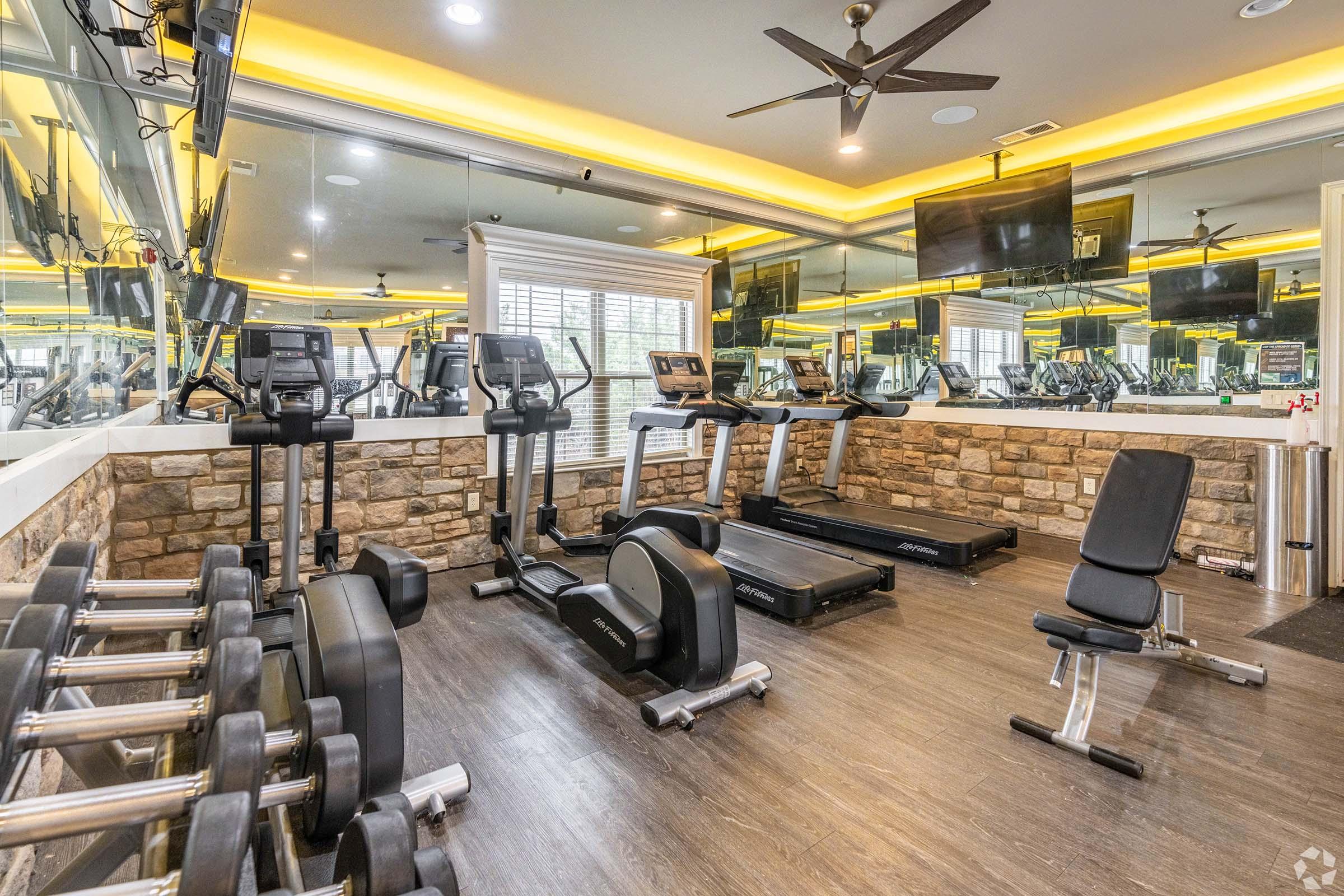 A modern gym area featuring cardio machines like treadmills and ellipticals, weight benches, and dumbbells. The space has a sleek design with stone walls, large mirrors, and bright ceiling lights, creating an inviting atmosphere for fitness activities. Two televisions are mounted on the walls for entertainment.