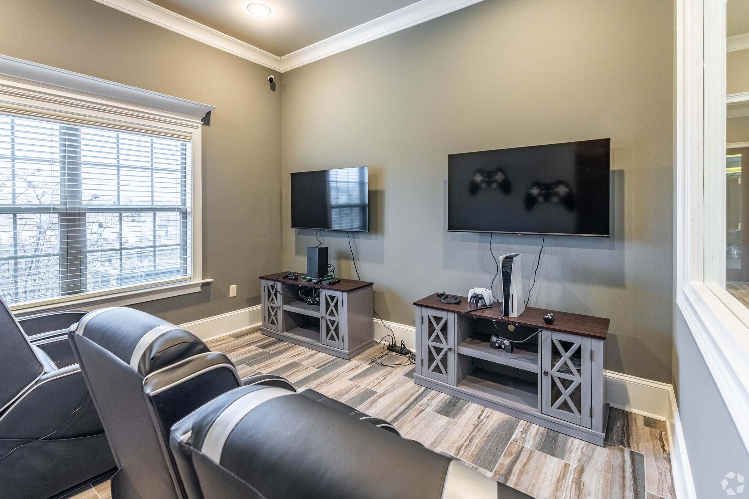 A cozy entertainment room featuring two wall-mounted flat-screen TVs, a gaming console, and a modern media cabinet. The space has comfortable black recliner chairs and large windows with horizontal blinds, allowing natural light to fill the room. The flooring is a light wood-like tile.