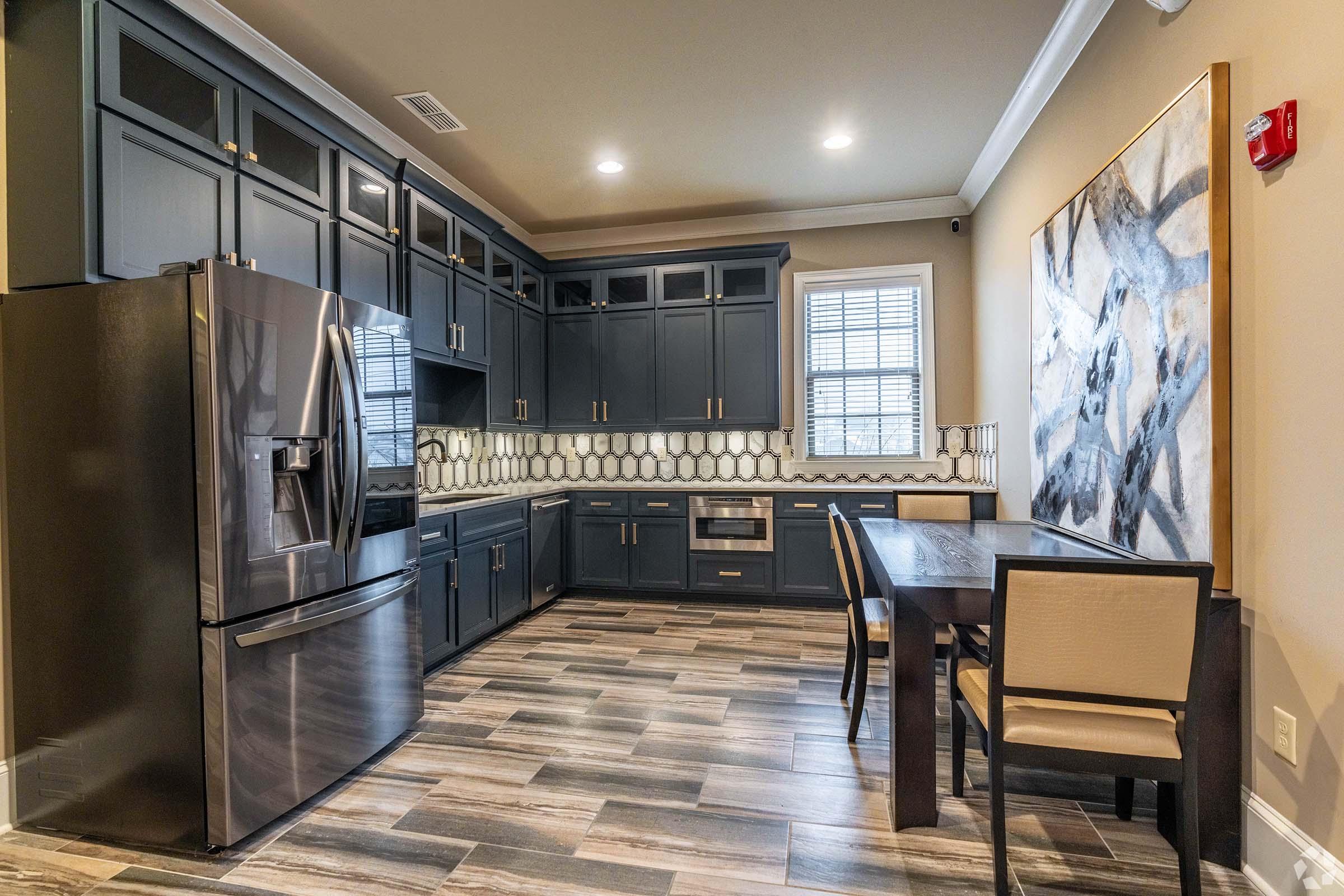 Modern kitchen featuring dark cabinetry, stainless steel refrigerator, and a compact dining area. The room includes a geometric backsplash, large window for natural light, and stylish wooden flooring. A wall art piece adds a contemporary touch to the space.