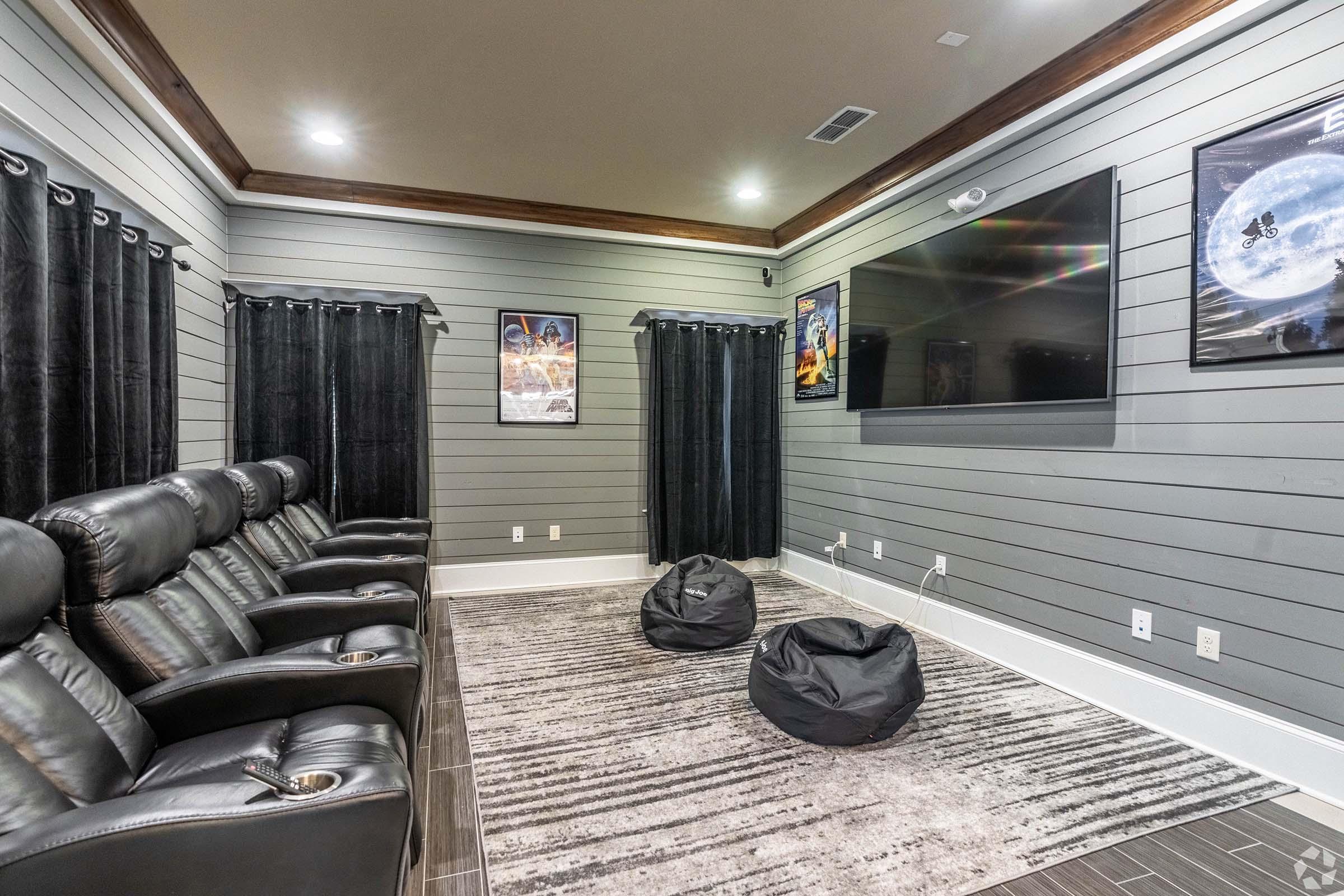 A cozy home theater featuring black reclining chairs with cup holders, bean bags for seating, dark curtains, and gray wood paneling. A large flat-screen TV is mounted on the wall, complemented by movie posters adorned around the room, creating an inviting cinematic environment.