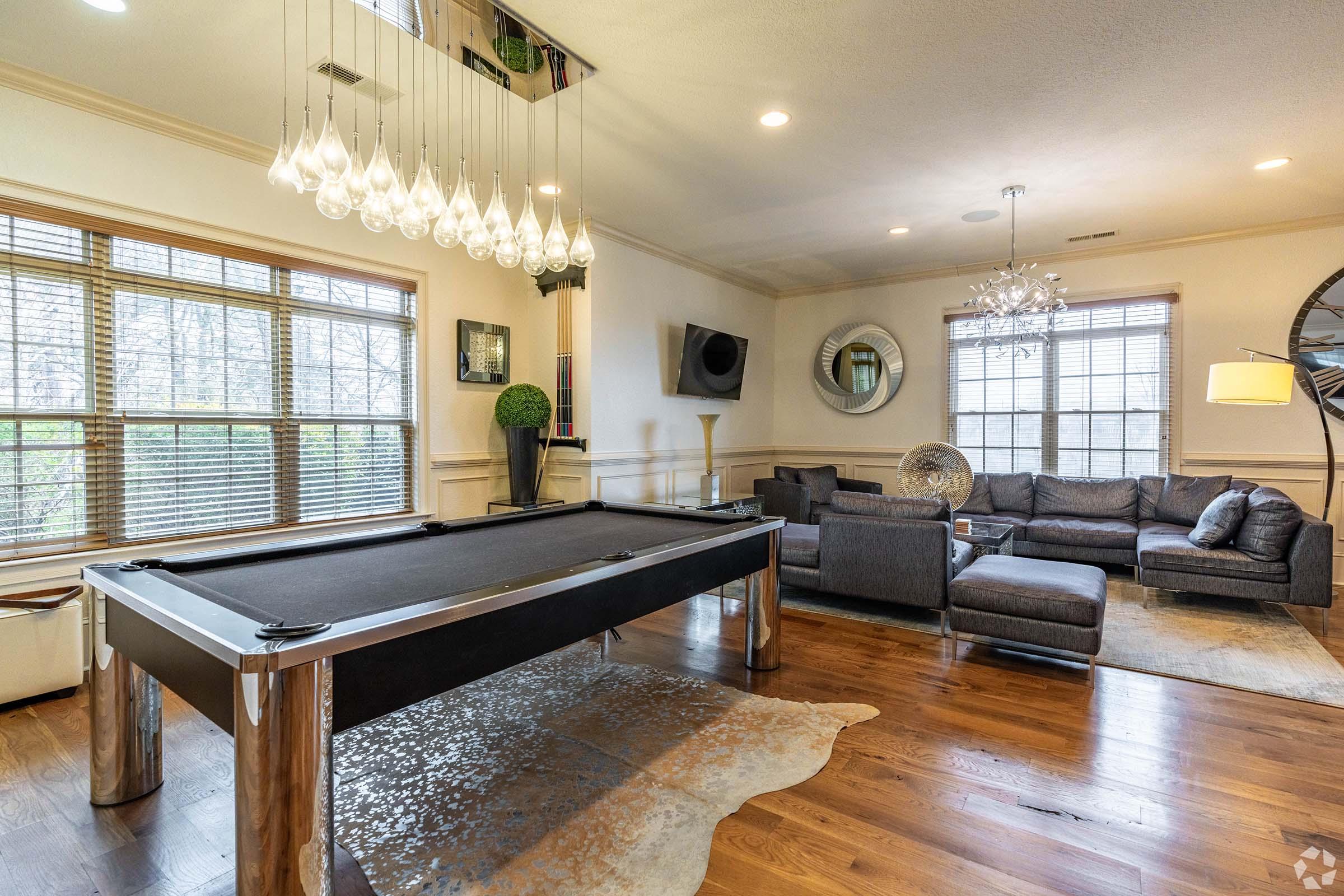 A stylish living room featuring a pool table, modern chandelier, comfortable gray seating, and large windows with natural light. Decorative plants and a mirror add to the contemporary aesthetic, while the wooden floor and stylish accessories create a warm, inviting atmosphere.