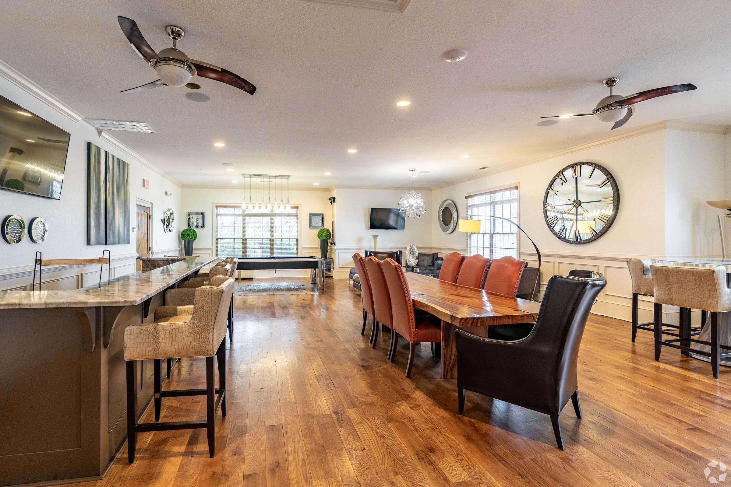 A spacious, modern living area featuring a large wooden dining table with multiple chairs, a stylish bar with stools, and a comfortable lounge area. The room is well-lit with natural light, has polished wooden floors, decorative wall art, and ceiling fans, creating an inviting atmosphere. Large windows offer views of the outdoors.