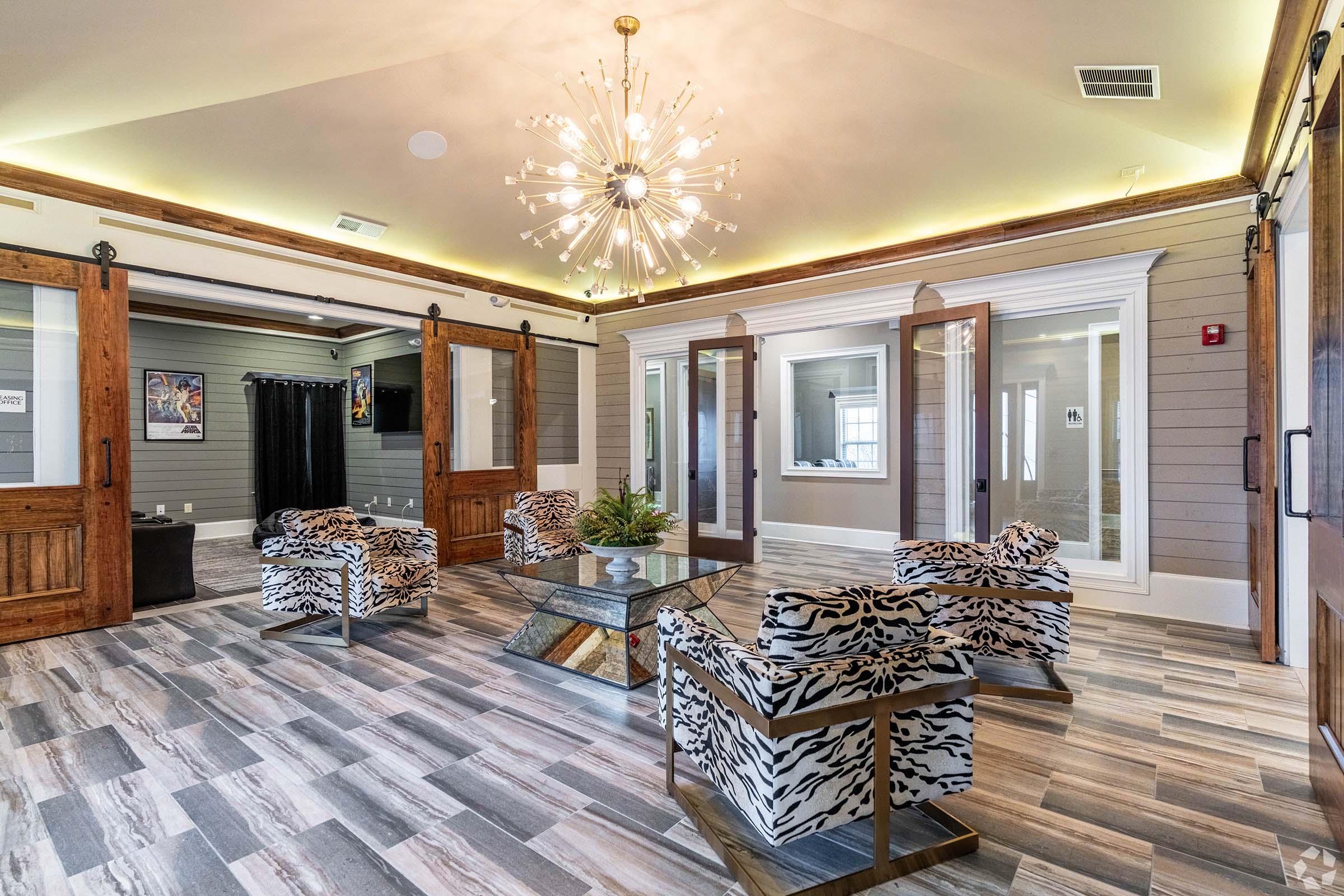 A stylish interior space featuring a decorative chandelier, modern zebra-print chairs, and a coffee table. Large glass doors lead to other areas, with light wood accents and patterned flooring. The walls are painted in muted tones, creating a contemporary and inviting atmosphere.