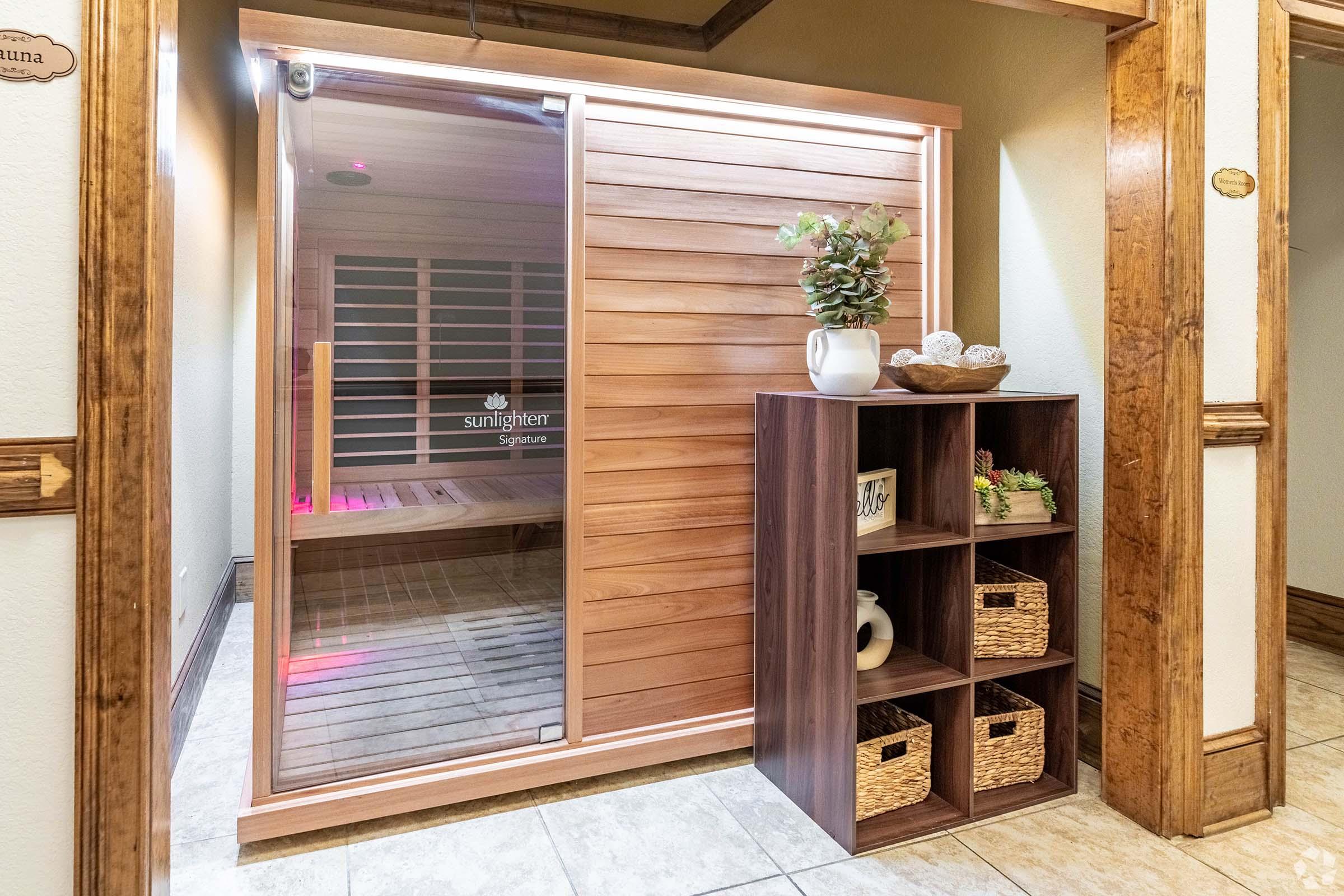 A modern sauna with wooden paneling, featuring a glass door. Next to it, there is a decorative shelf unit with woven baskets and a plant. The surroundings have a warm, inviting atmosphere, highlighted by soft lighting.